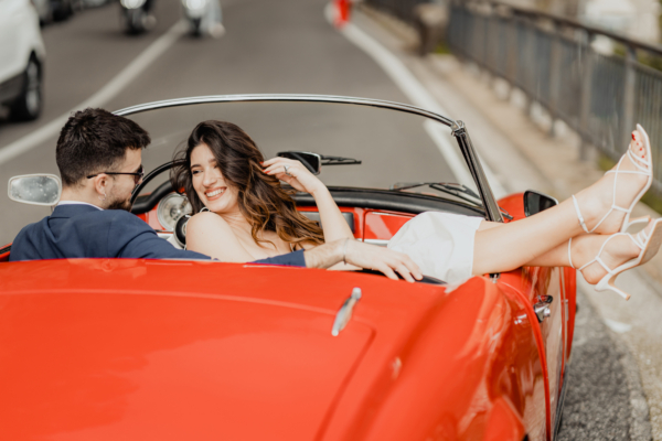 Bride and groom in vintage red car laughing together - cinematic destination wedding photography in Italy by Calini Weddings.