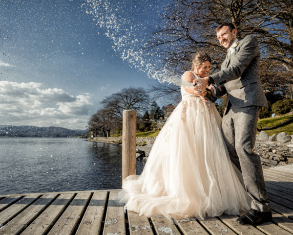 Bride and groom celebrating with champagne by the lake - joyful Lake District wedding photography by Calini Weddings.