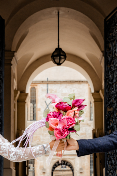 Vibrant pink and coral bridal bouquet held between couple under elegant arch - modern wedding photography by Calini Weddings.
