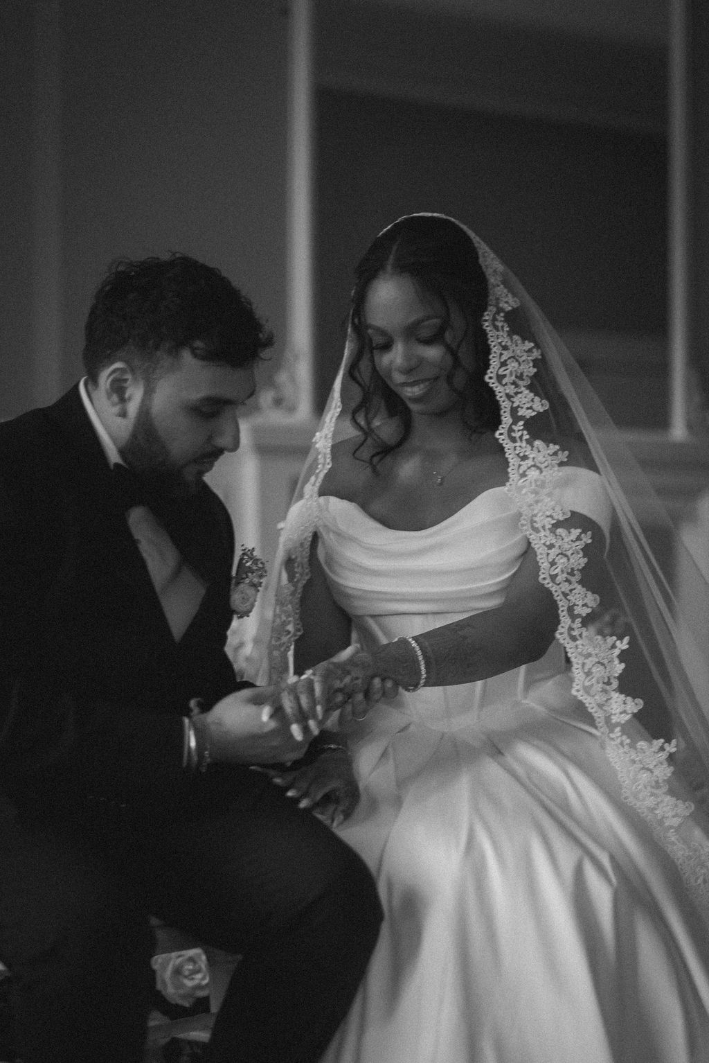 Black bride wearing an off the shoulder, corseted dress and mantilla lace veil, her groom gently holds her hands to observe her henna tattoo.