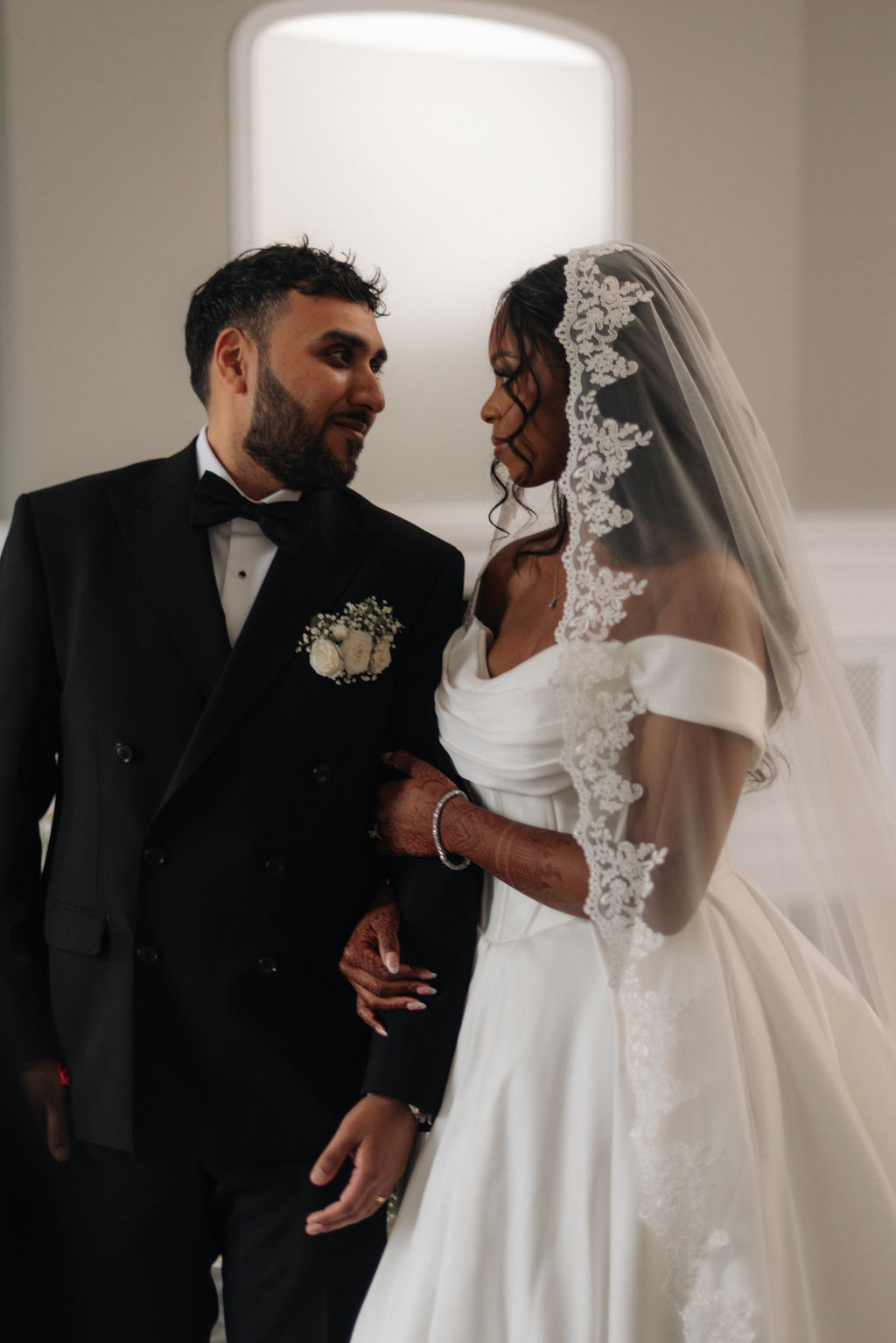 Bride in mantilla lace veil gently clutching the arm of her groom in black tie.