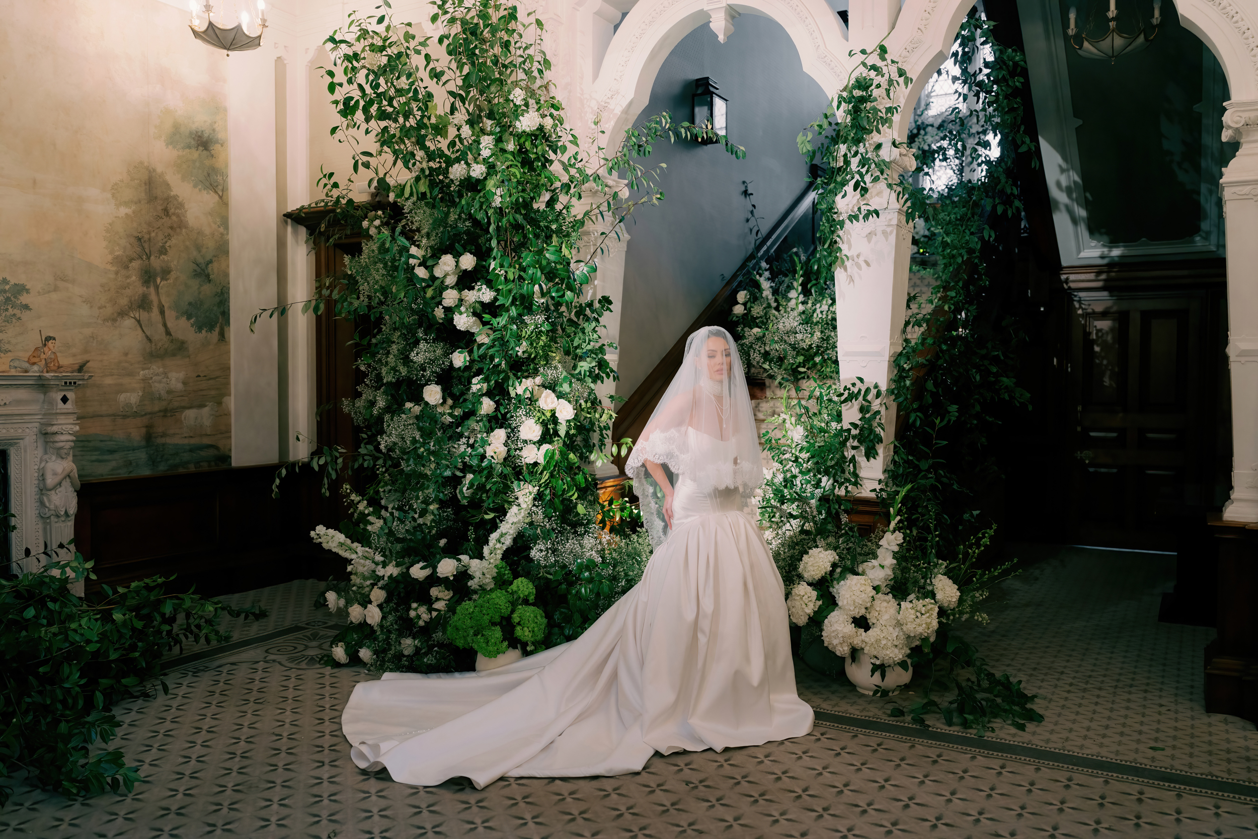 Bride standing by the staircase at Clevedon Hall wedding venue - an abundance of white roses and foliage surrounds her. She wears Josephine Scott London.