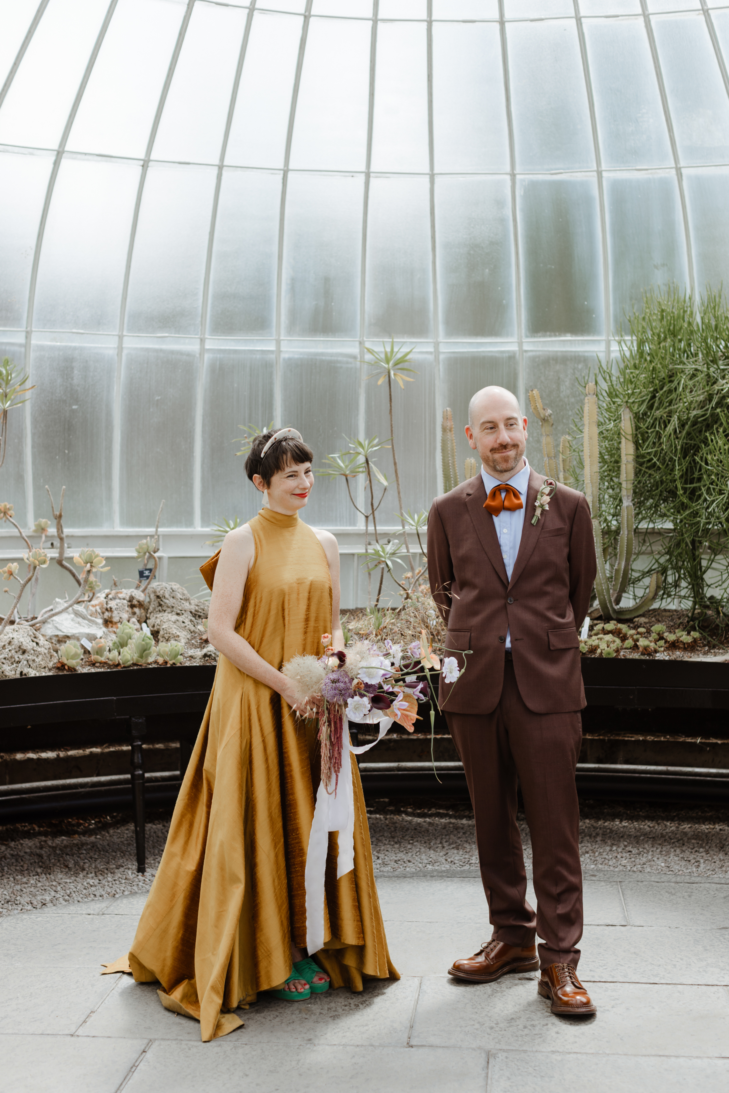 Chris and Barbara’s Delightful Family Wedding at Kibble Palace, Glasgow 18 Bride in a non traditional mustard yellow dress with large bow at the back. She wears green platform open toe shoes and carries a bouquet she made herself. Groom in a brown suit and draped silk ribbon bow tie.