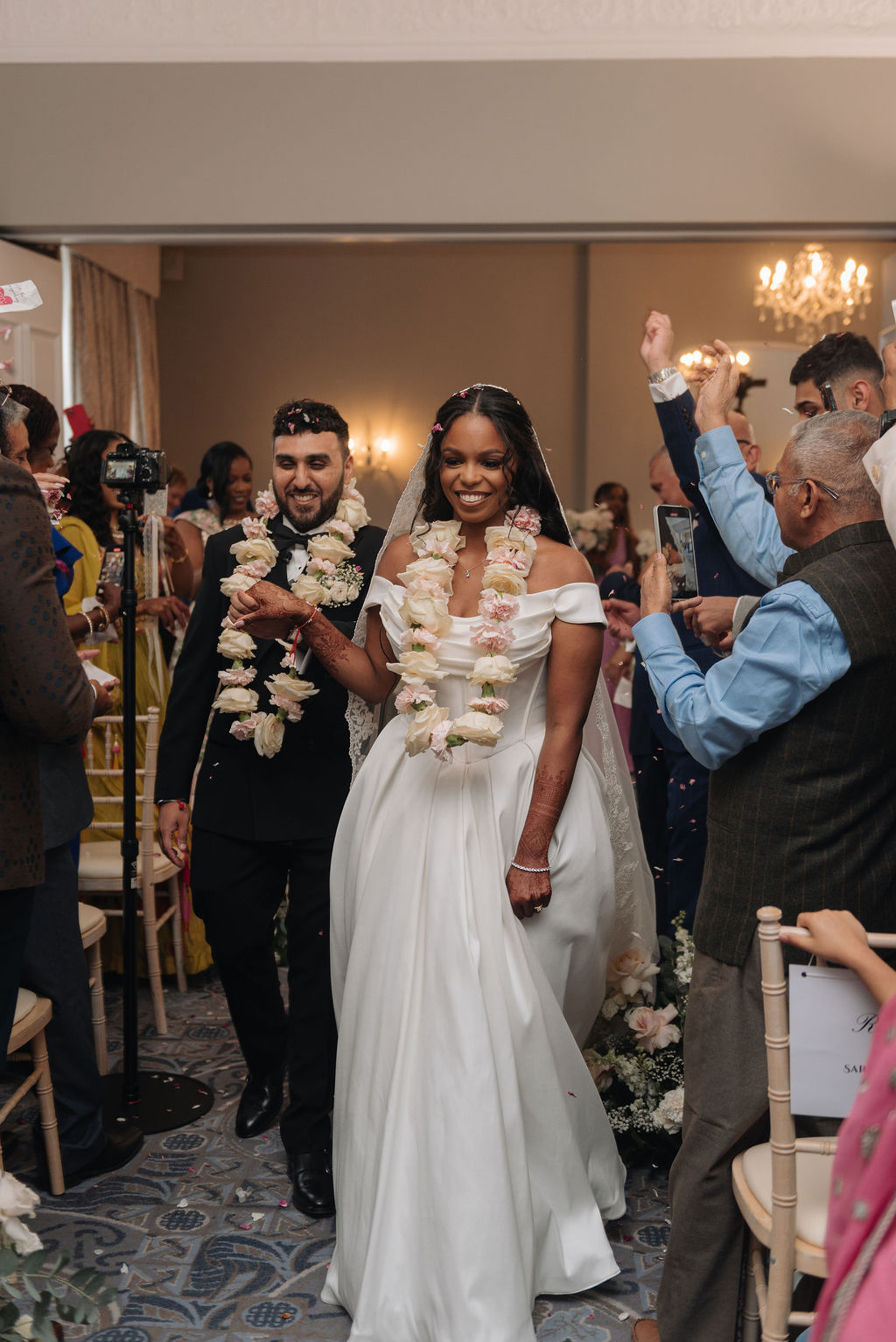 Bride and groom newlywed leaving their multicultural wedding ceremony at Oakley Hall. they both wear pale pink and white floral garlands around their necks.
