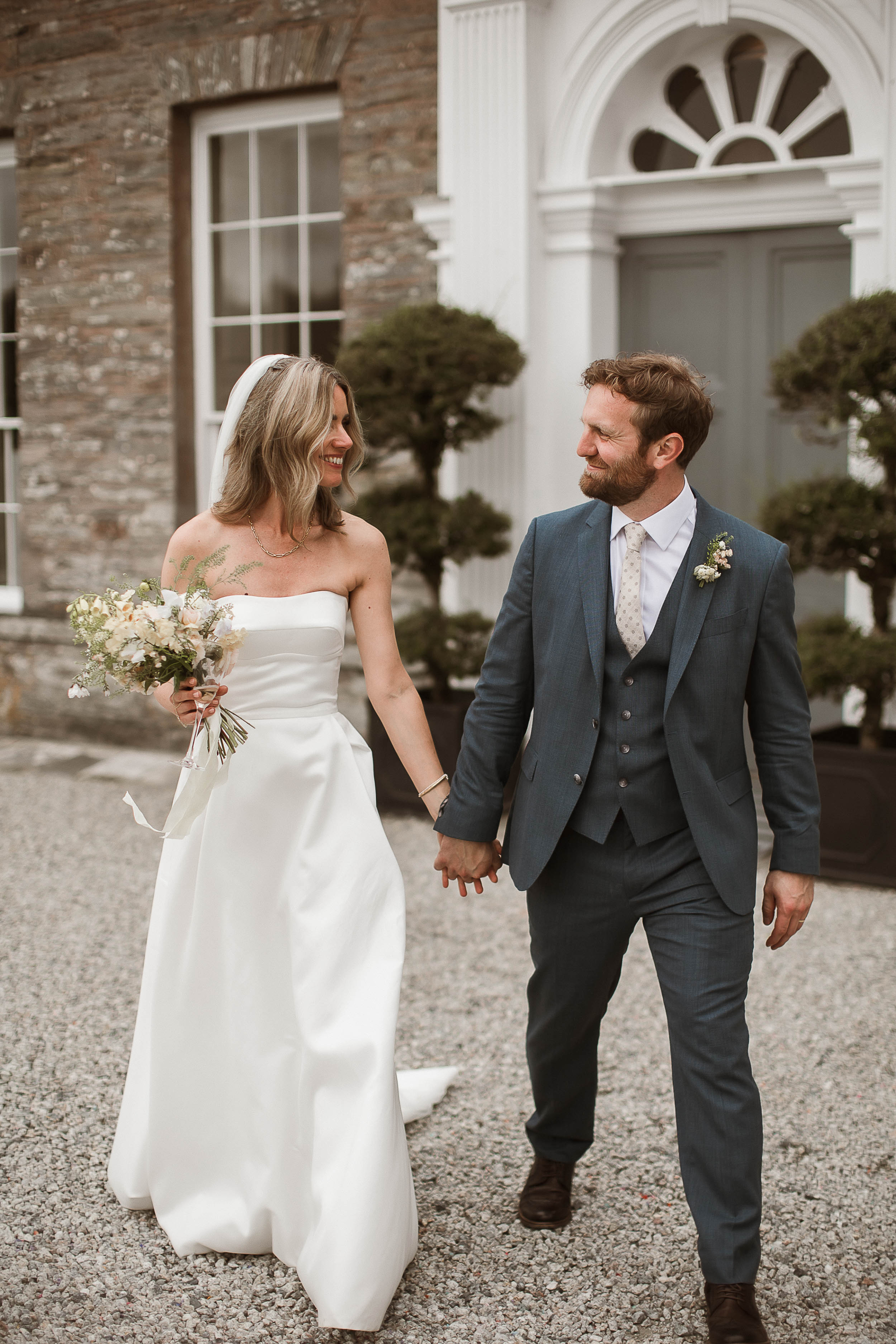 Boconnoc Estate Wedding Cornwall Jesus Peiro 137 - Lauren in Jesus Peiro for her Elegant Boconnoc Estate Wedding in Cornwall Bride with loose long blond hair, wearing a Jesus Peiro wedding dress clutching her groom's hand and holding an elegant summer wedding bouquet with the other. He wears a pale grey suit.