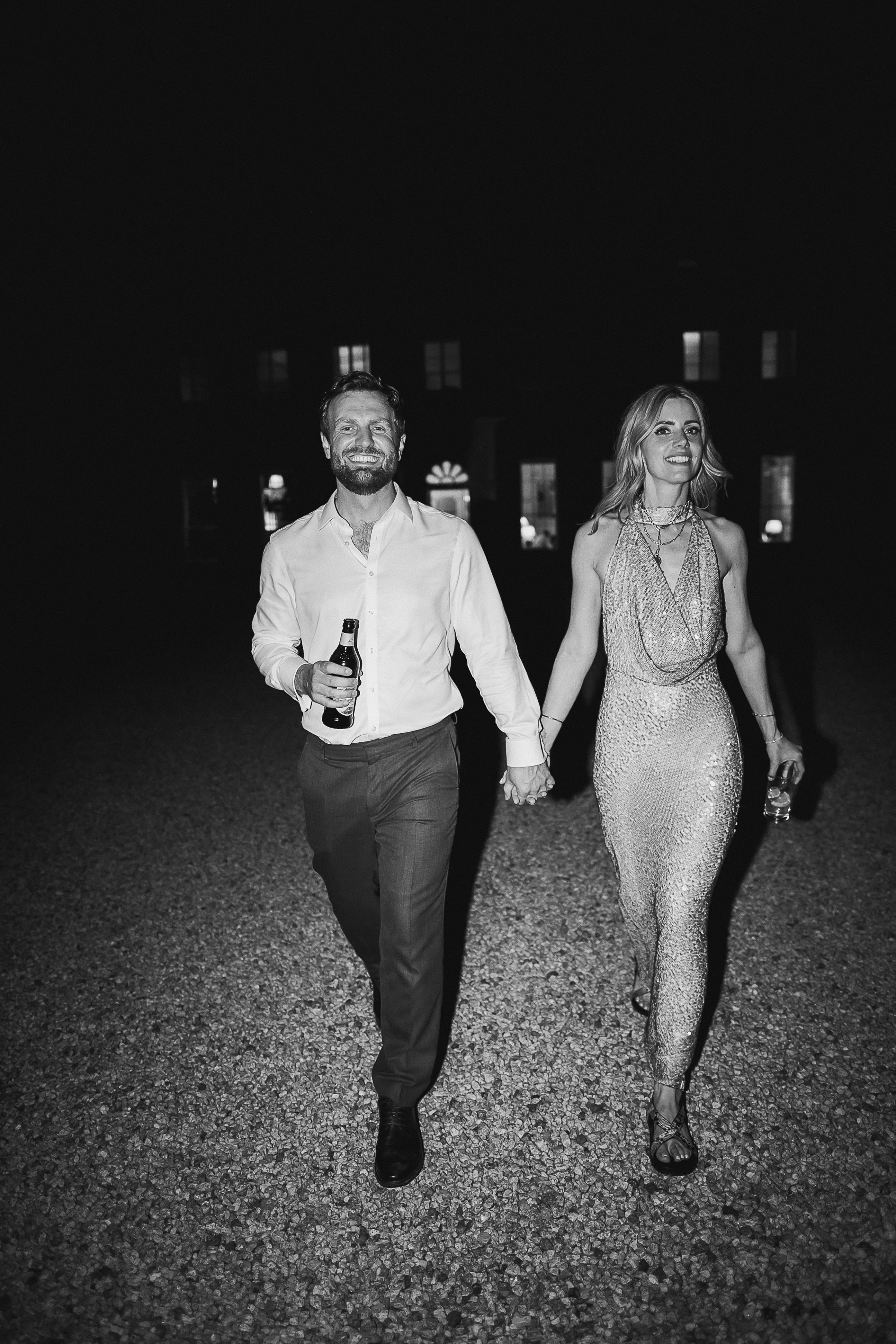 Boconnoc Estate Wedding Cornwall Jesus Peiro 192 - Lauren in Jesus Peiro for her Elegant Boconnoc Estate Wedding in Cornwall Bride in a sequin halterneck dress for the evening of her wedding, holding hands with her groom who wears a shirt unbuttoned at the top.