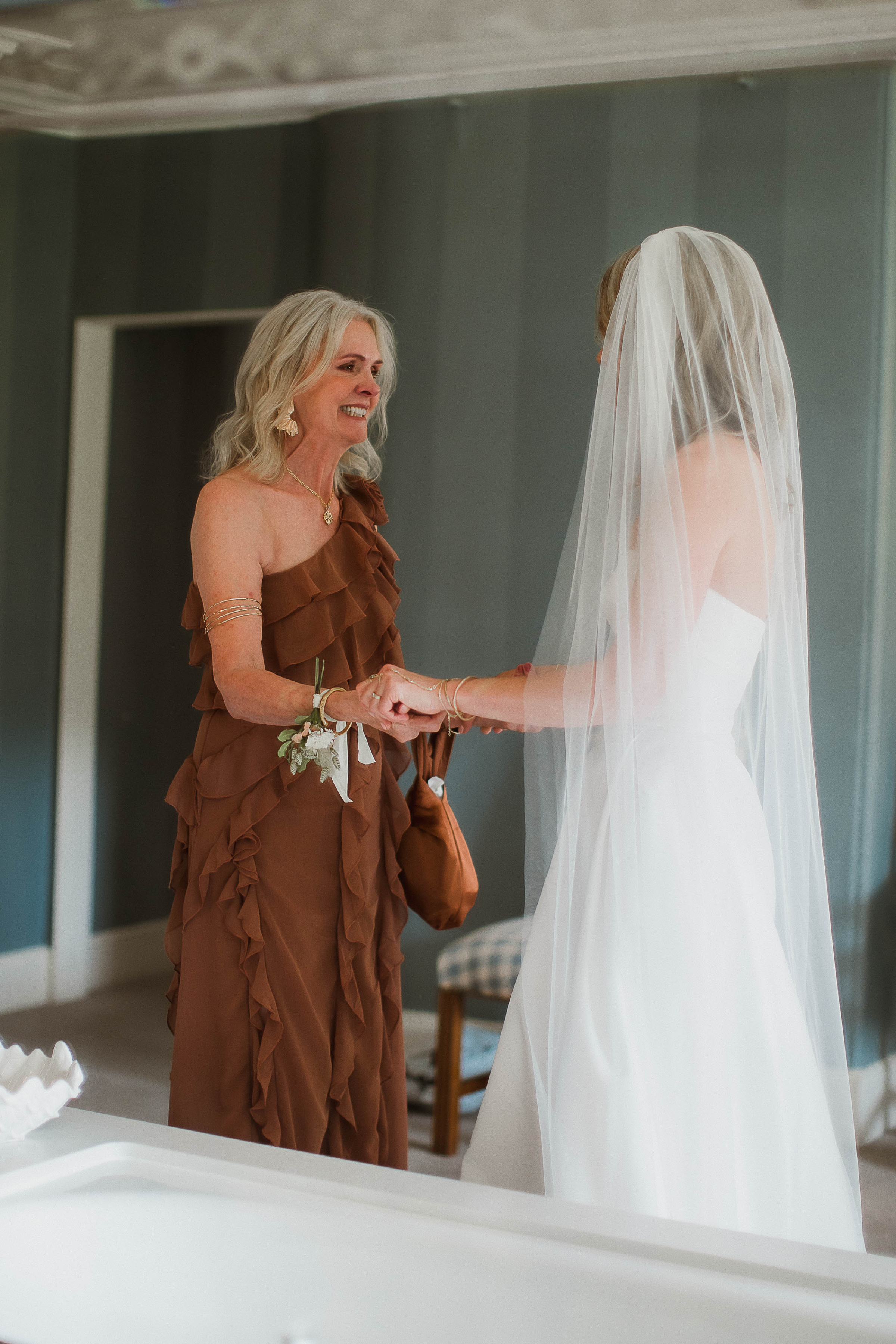 Boconnoc Estate Wedding Cornwall Jesus Peiro 45 - Lauren in Jesus Peiro for her Elegant Boconnoc Estate Wedding in Cornwall Mother of the bride in an asymmetrical brown dress clutching the hands of her daughter as she smiles at her. We see the back of the bride as she looks on at her mum.