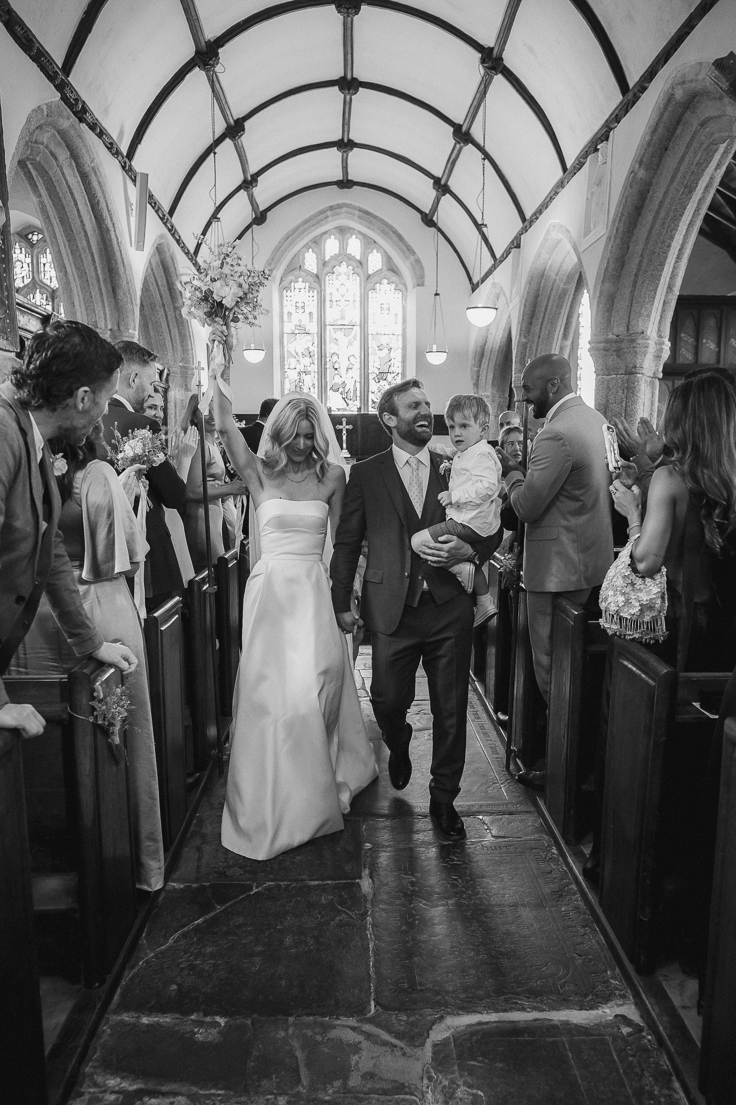 Boconnoc Estate Wedding Cornwall Jesus Peiro 73 - Lauren in Jesus Peiro for her Elegant Boconnoc Estate Wedding in Cornwall Bride and groom exiting their church wedding ceremony. The groom has a child in one arm and the bride is raising her arm and bouquet into the air.