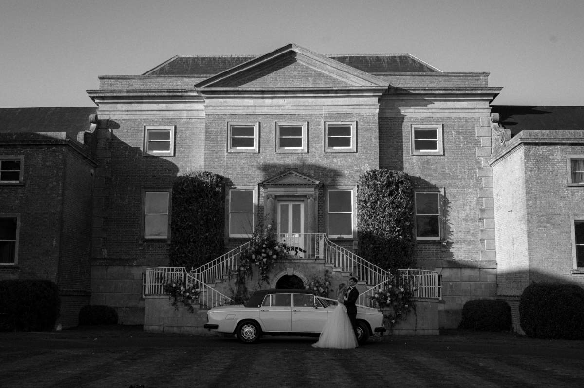 Earnshill350 - Modern Bridal Elegance at Earnshill House, A Romantic Somerset Wedding Venue Vintage car parked outside Earnshill House, A Romantic Somerset Wedding Venue. Bride and groom standing nearby and embracing by the car.