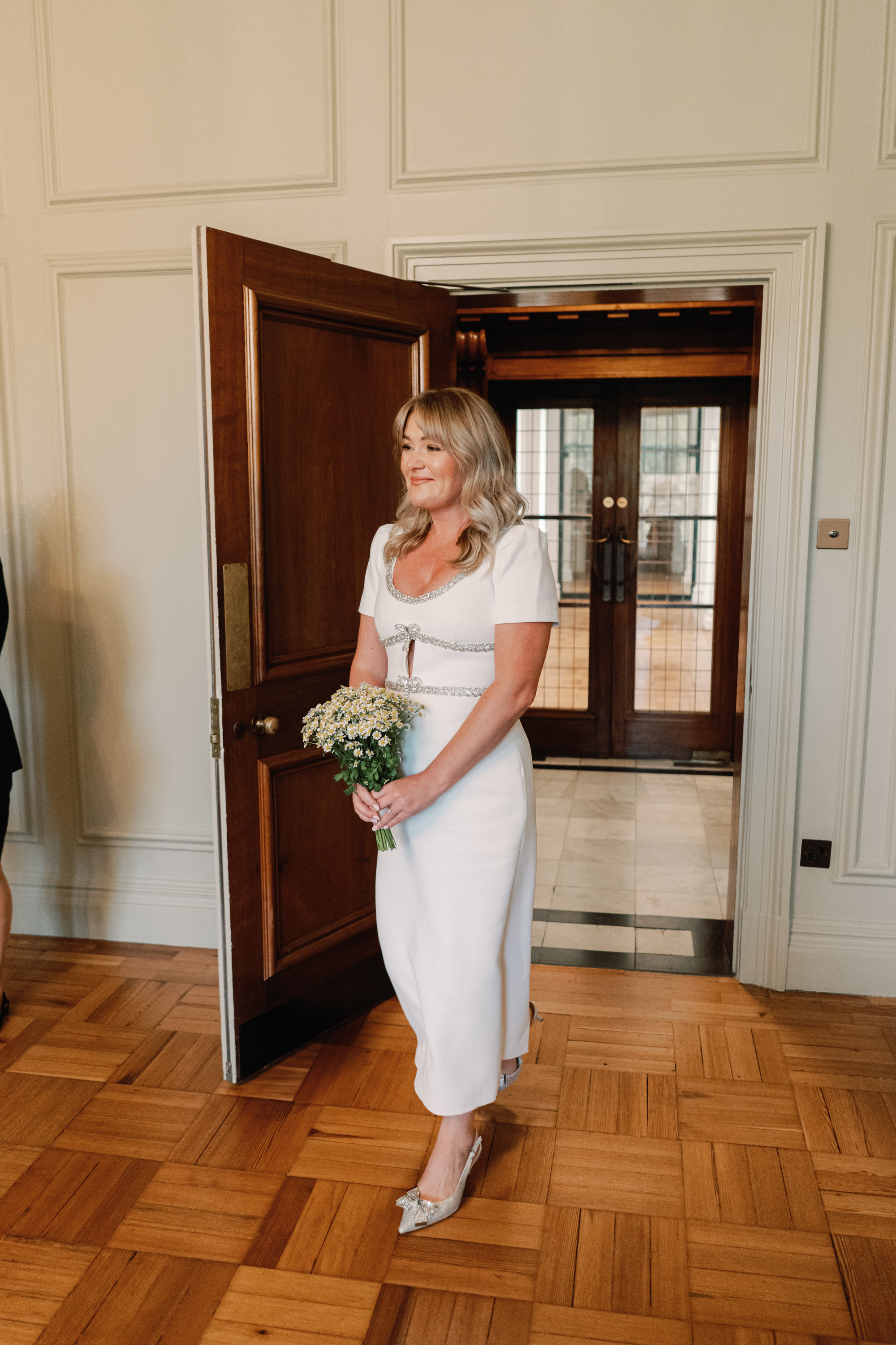 Emmylou Kelly Registry Office Wedding Jessica Lily Photography 103 - Photographer Emmylou Kelly & Paul's Intimate Family Registry Office Wedding Bride in a midi wedding dress by Self Portrait entering the ceremony room at Old Marylebone Town Hall. She carries a small bouquet of Chamomile.
