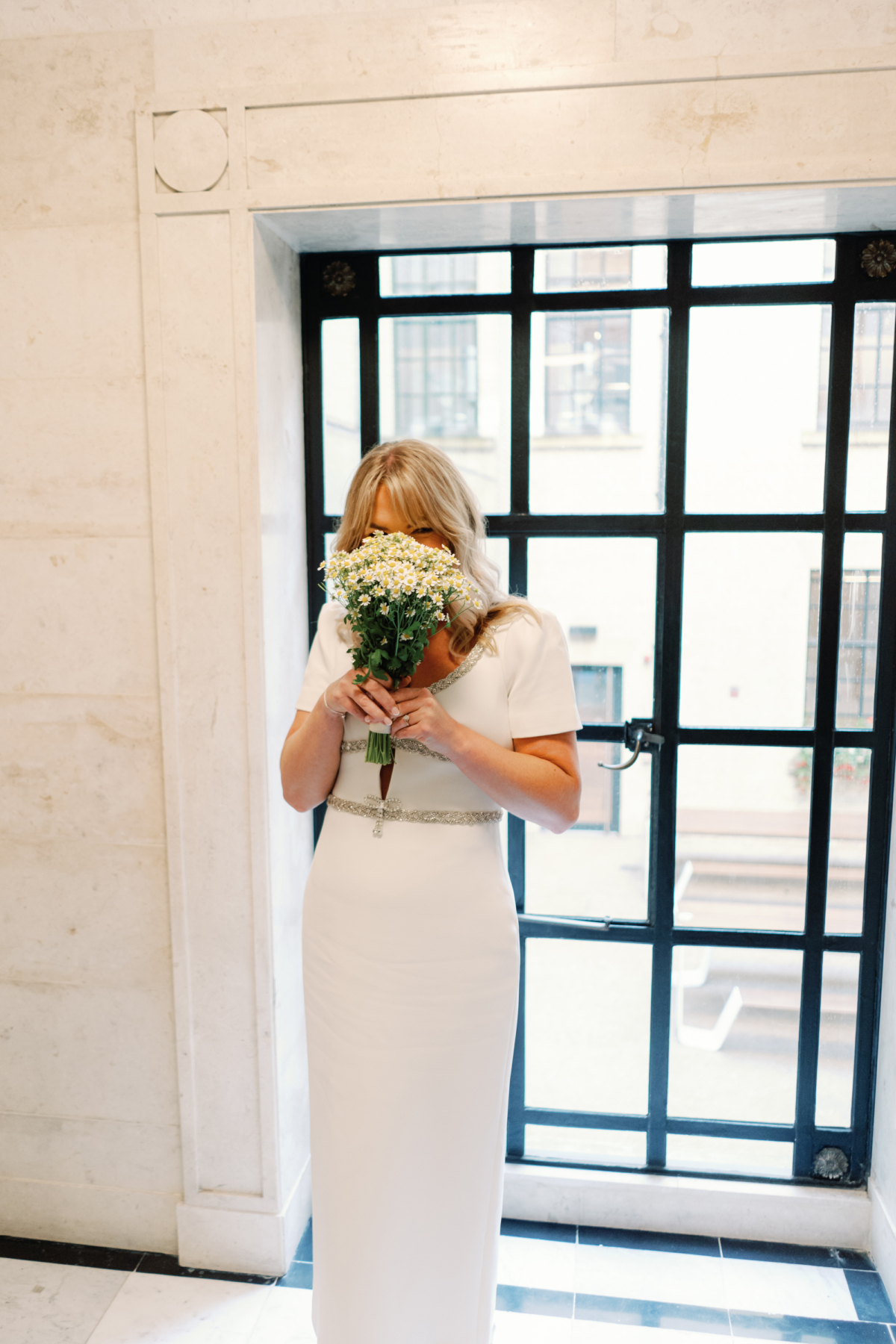 Bride hiding her face behind her bouquet of Chamomile. She wears a midi dress by Self Portrait.