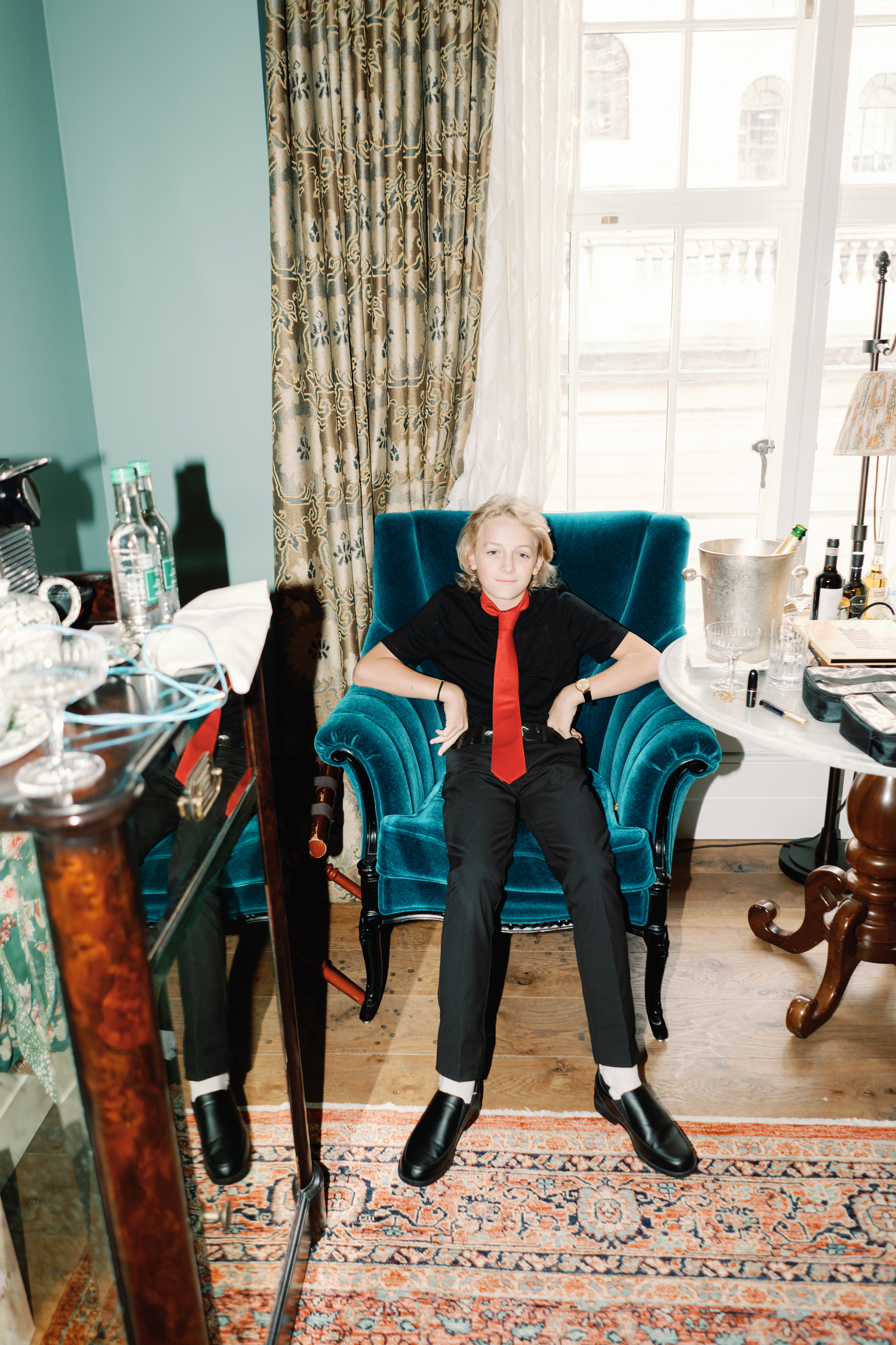 Emmylou Kelly Registry Office Wedding Jessica Lily Photography 77 - Photographer Emmylou Kelly & Paul's Intimate Family Registry Office Wedding Young male wedding guest sat on a turquoise velvet chair. He wears black trousers and shirt with a red tie.
