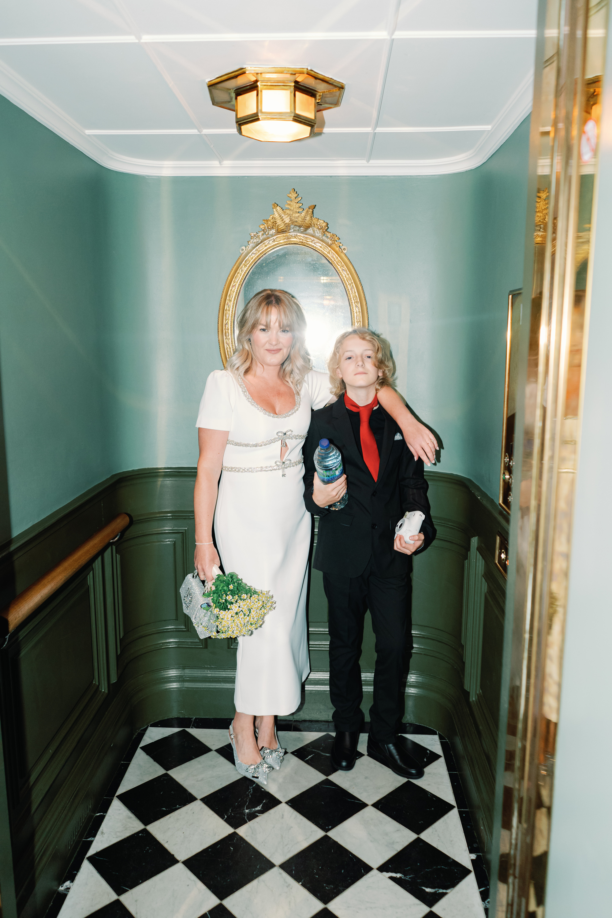 Emmylou Kelly Registry Office Wedding Jessica Lily Photography 95 - Photographer Emmylou Kelly & Paul's Intimate Family Registry Office Wedding Bride standing in a lift with chequered flooring. She wears a midi dress by Self Portrait, sparkly bow shoes by Carvela and carries a small chamomile bouquet. She has her arm around her son in black trousers and shirt and a red tie.