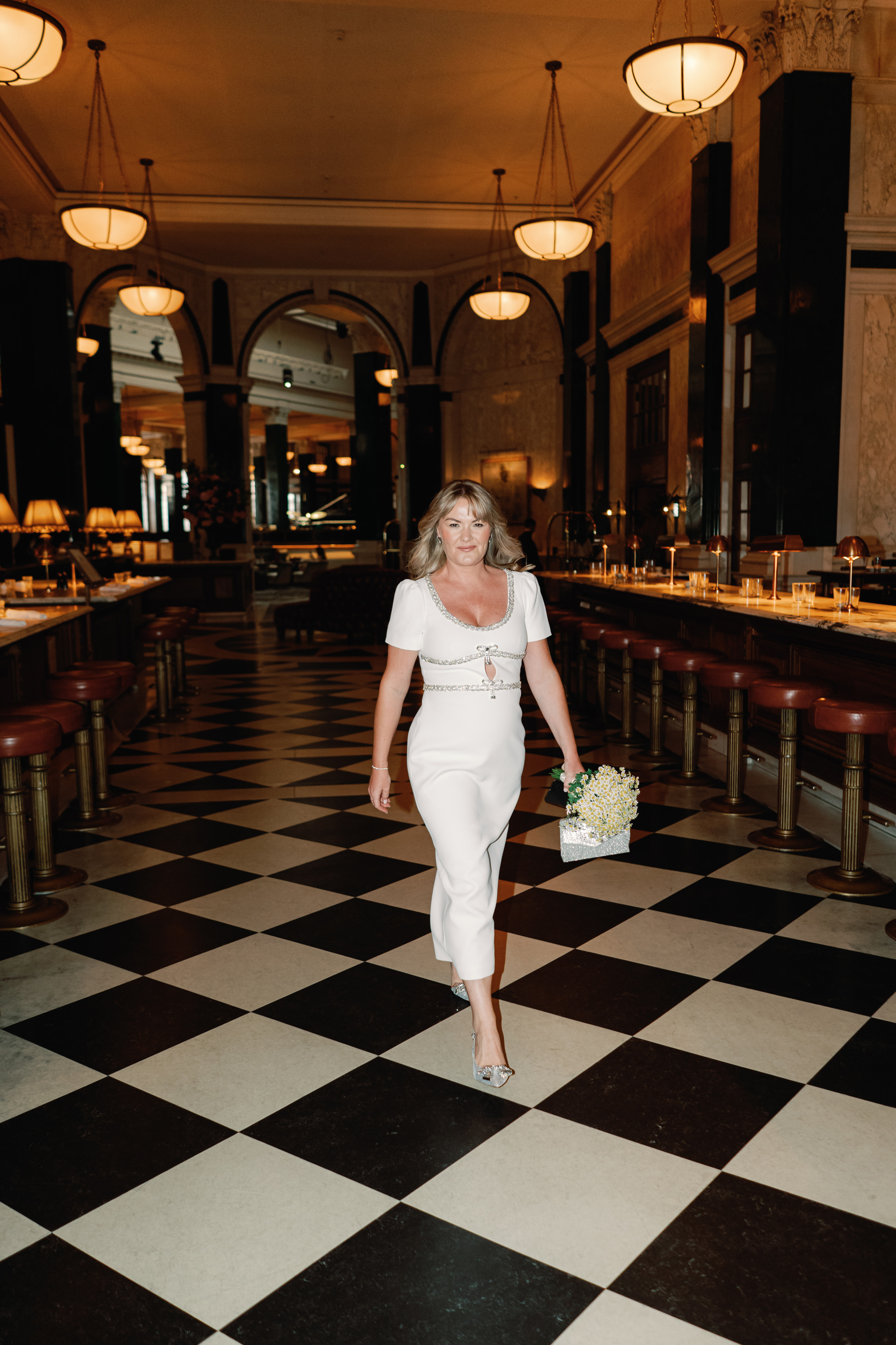 Brirde wearing a midi dress by Self Portrait and sparkly bow shoes by Carvela walking across a black and white chequered floor at The Ned Hotel in London . She carries a small chamomile bouquet and clutch bag.