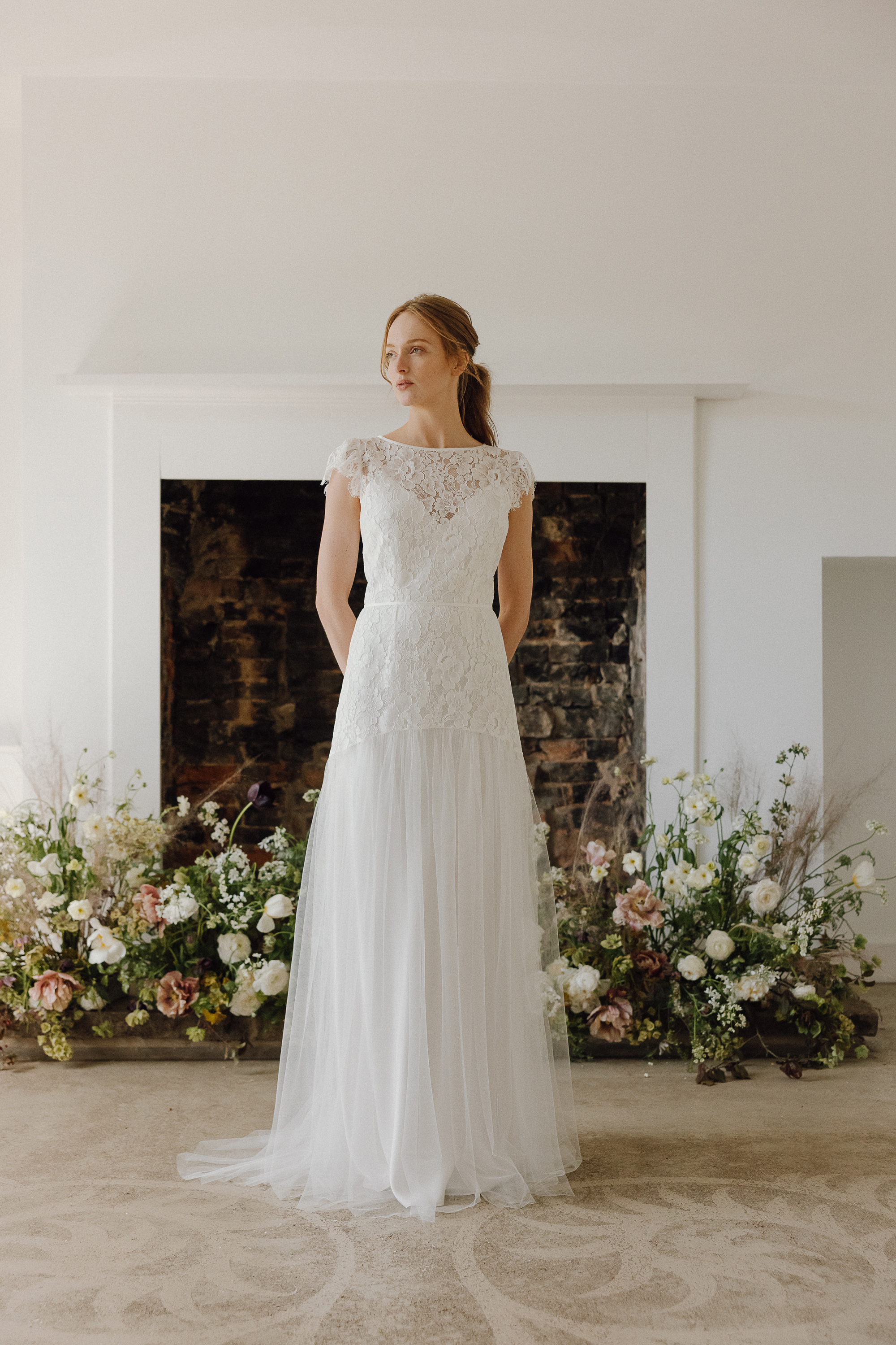 Geum lace wedding gown KateBeaumont 2025 IndiaHobson HR 339 1 - Kate Beaumont: Lovingly Handmade Silk Wedding Dresses Elegant modern lace and tulle wedding dress by Kate Beaumont. Model bride stands in front of a fireplace decorated with beautiful flowers by Swallows & Damsons.