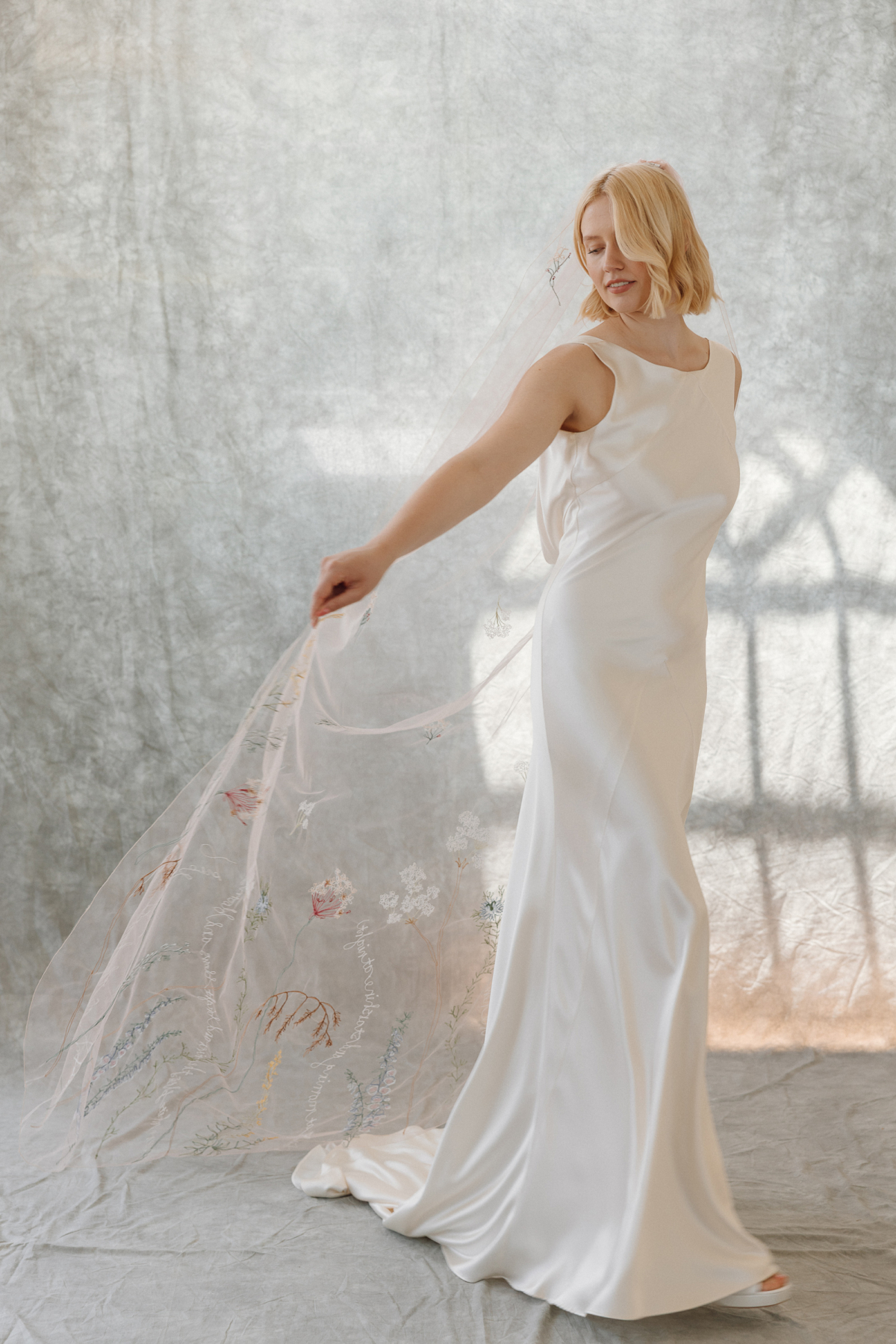 Bride wearing a silk bias cut wedding dress by Kate Beaumont with an embroidered wedding veil by Daisy Sheldon, featuring colourful embroidered flowers, leaves and words.
