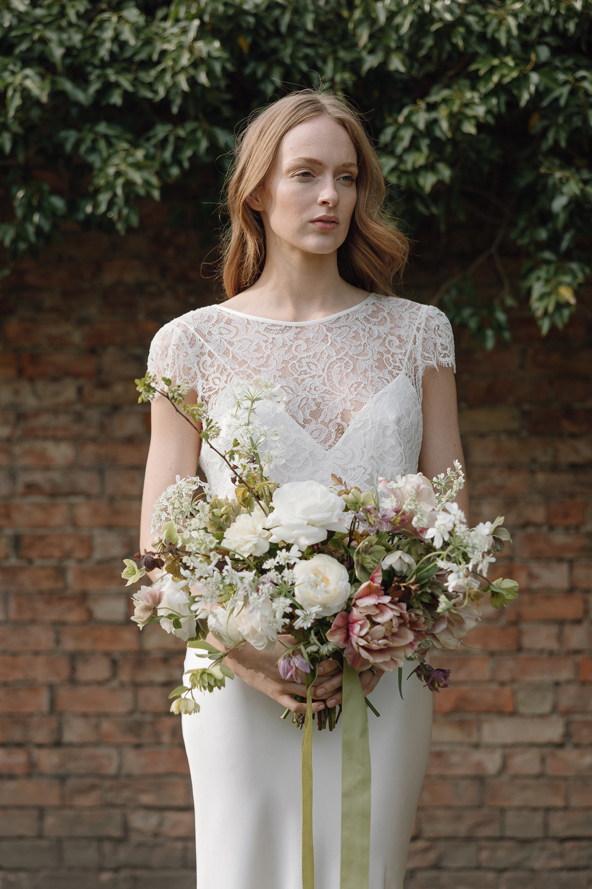Jasmine bridal lace top KateBeaumont 2025 IndiaHobson HR 611 - Kate Beaumont: Lovingly Handmade Silk Wedding Dresses Bride with minimalist silk slip dress and lace overlay by Kate Baumont, holding a n elegant bouquet of flowers in pale greens and pinks with long green silk ribbon.
