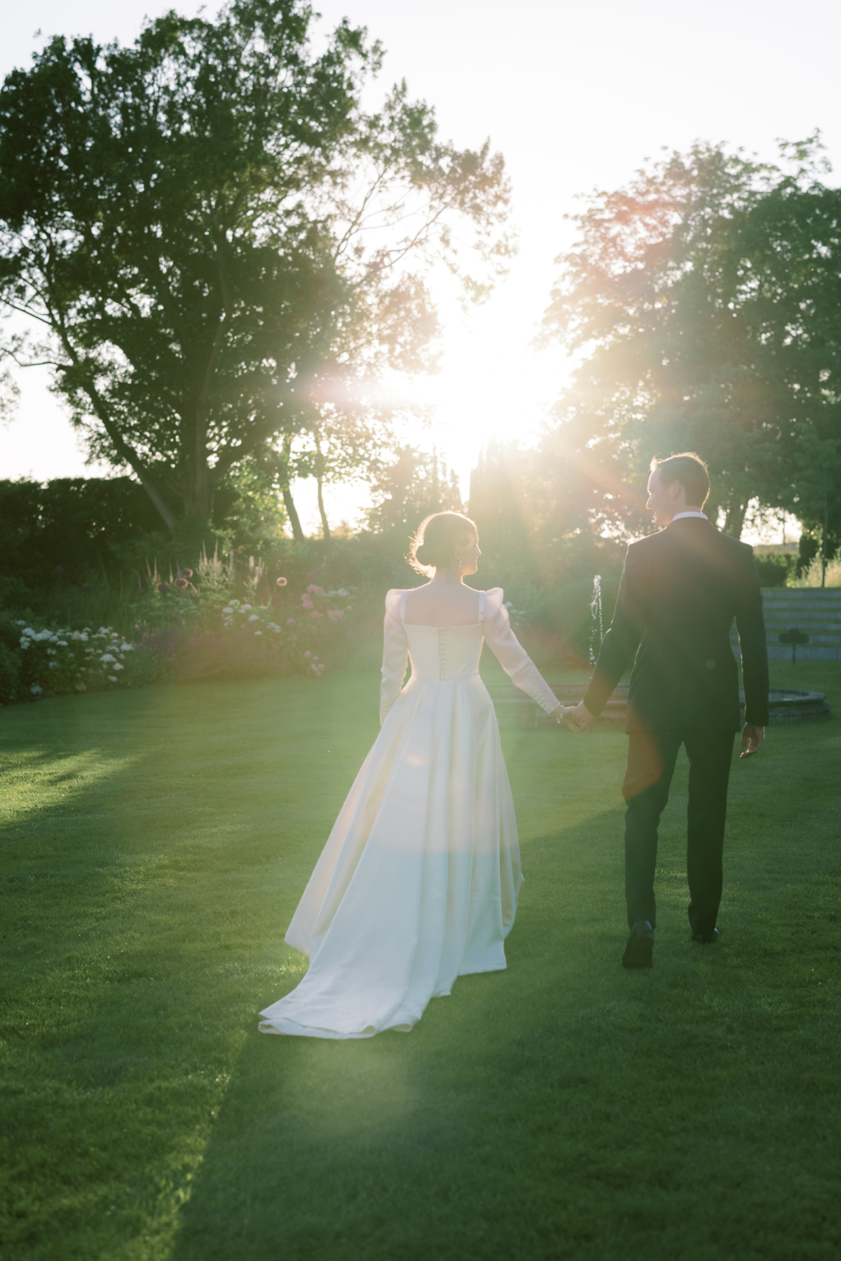 Golden hour shot of bride wearing a Phillipa Lepley wedding dress holding hands with her groom in black tie.