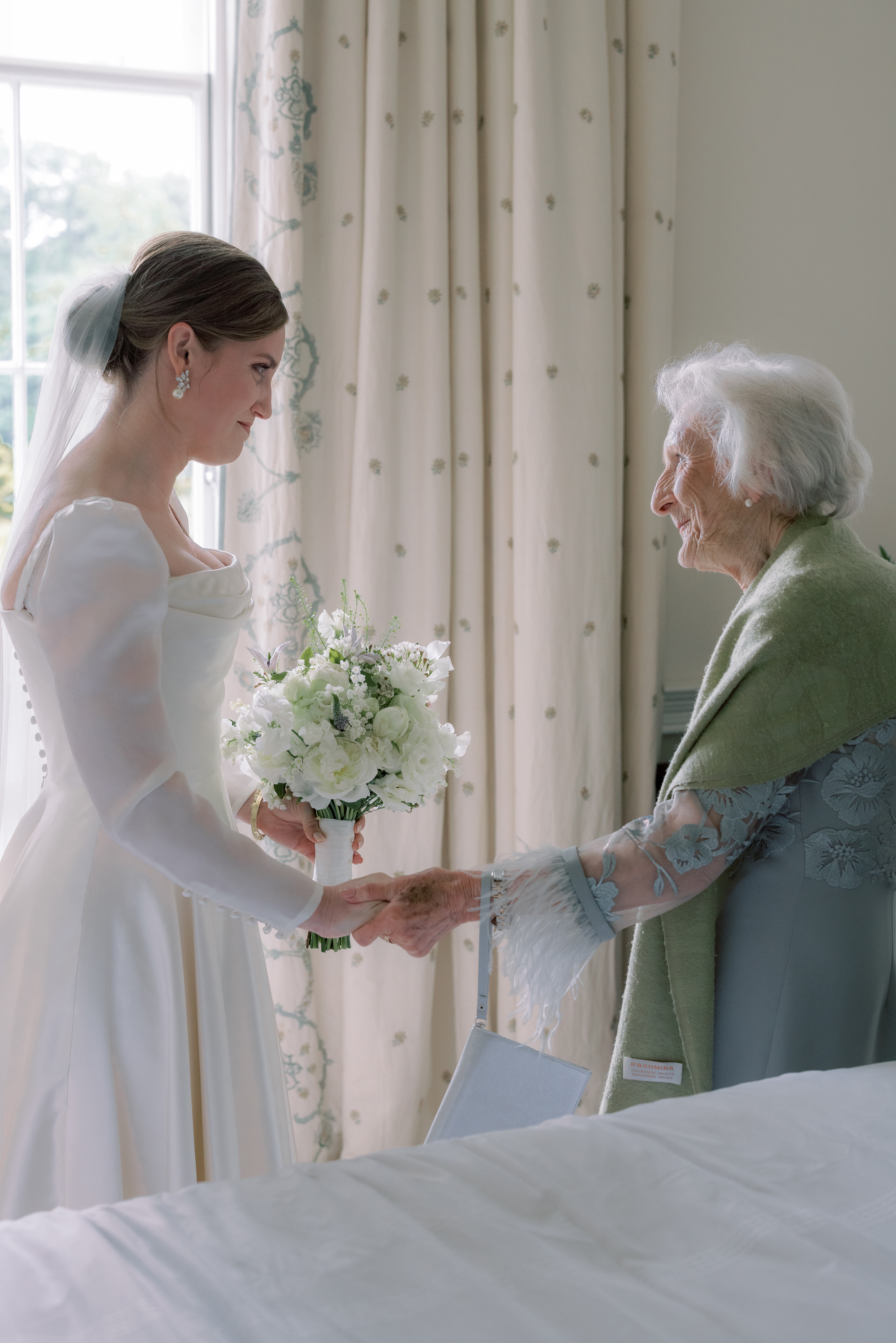 Tender wedding photograph of a bride in a Phillipa Lepley wedding dress holding the hands of her elderly relative.She also holds an elegant all-white wedding bouquet.