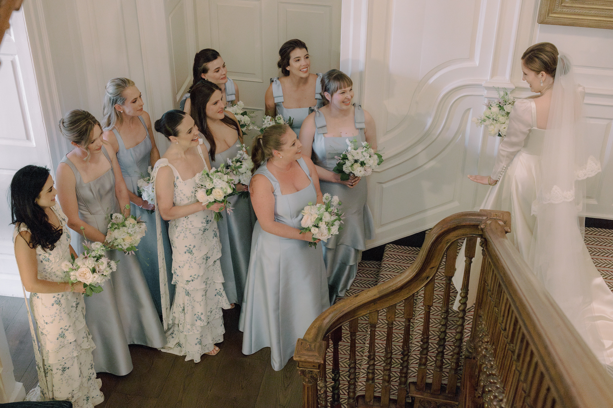Bridesmaids in pale blue gathered at the bottom of a sweeping staircase, looking up at the bride descending the stairs in her Phillipa Lepley wedding dress.