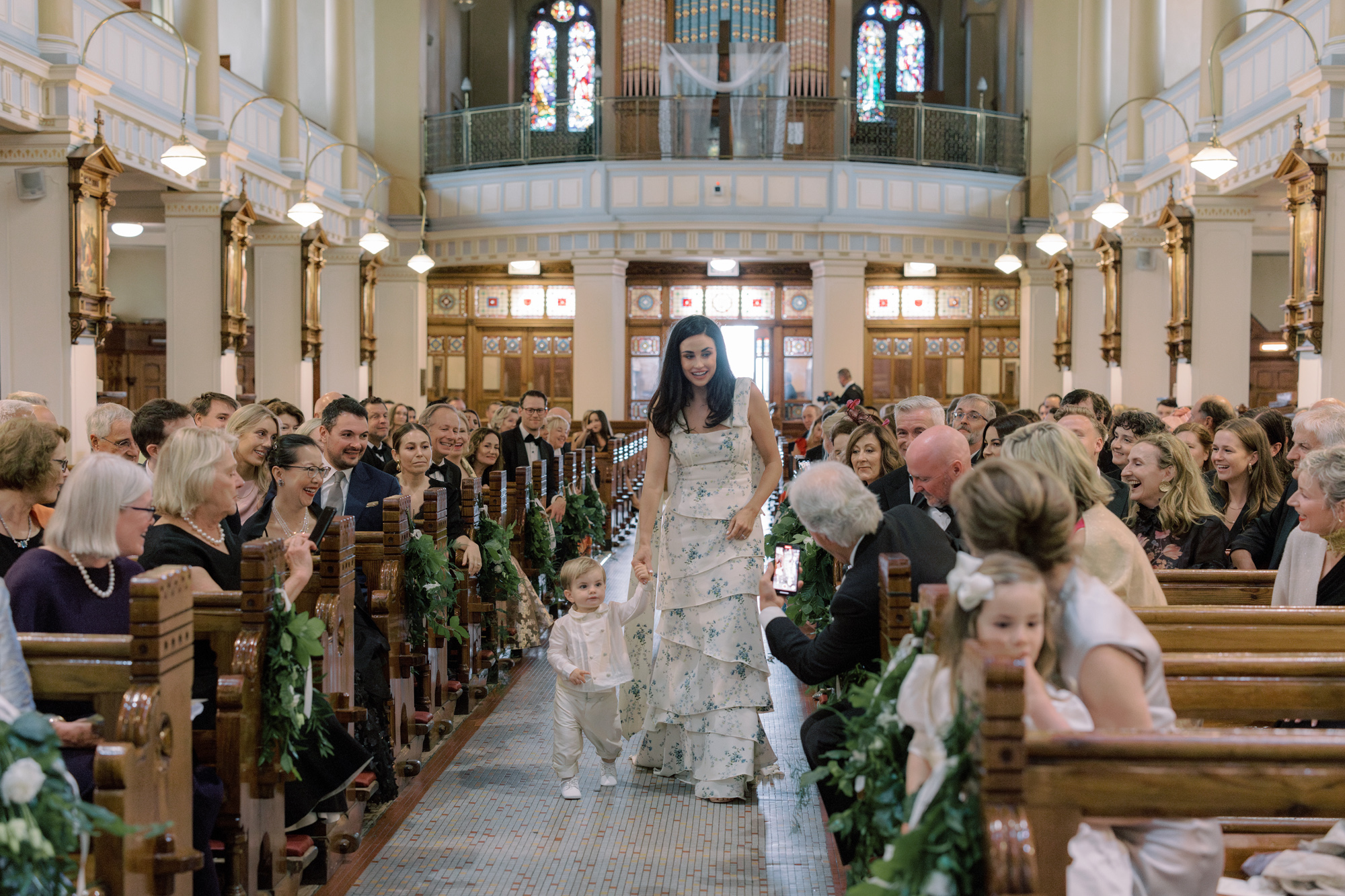 Bridesmaid in a pale blue flower print dress walking down hte aisle with a todler page boy holding her hand.