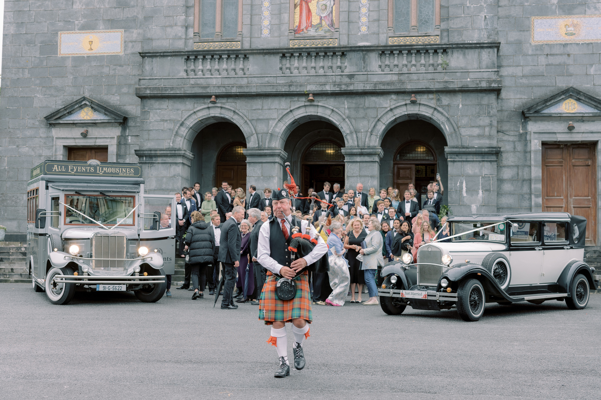 Bagpiper playing outdoors nearby a gathered wedding crowed who are standing next to a vintage car and bus.