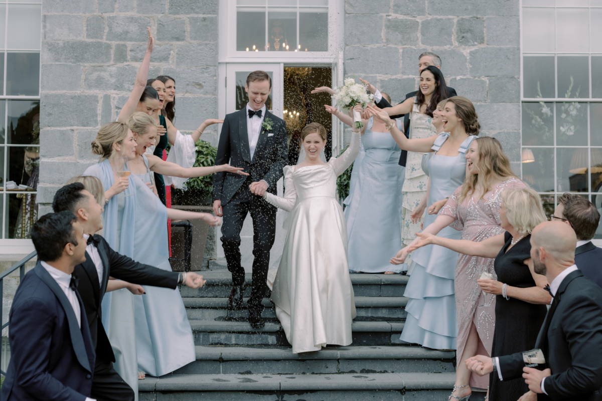 Bride wearing Phillipa Lepley walking hand in hand with her groom in black tie down a flight of steps as they are showered in confetti by bridesmaids in mismatched pale blue dresses.