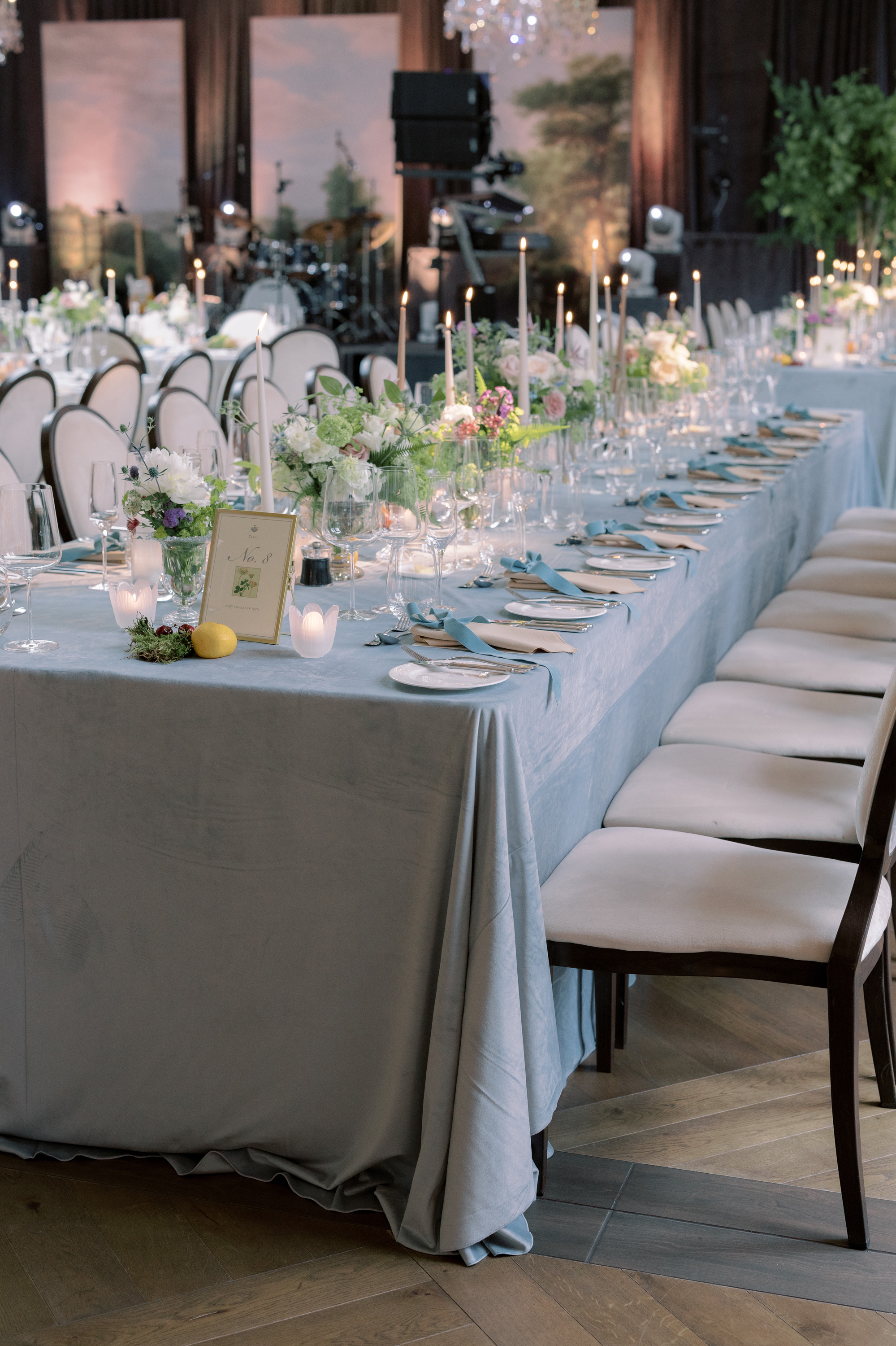 Long table at a wedding reception wiht a pale blue table cloth, pale blue embroidered ribbons used as place names, tapered candles and elegant low flowers.
