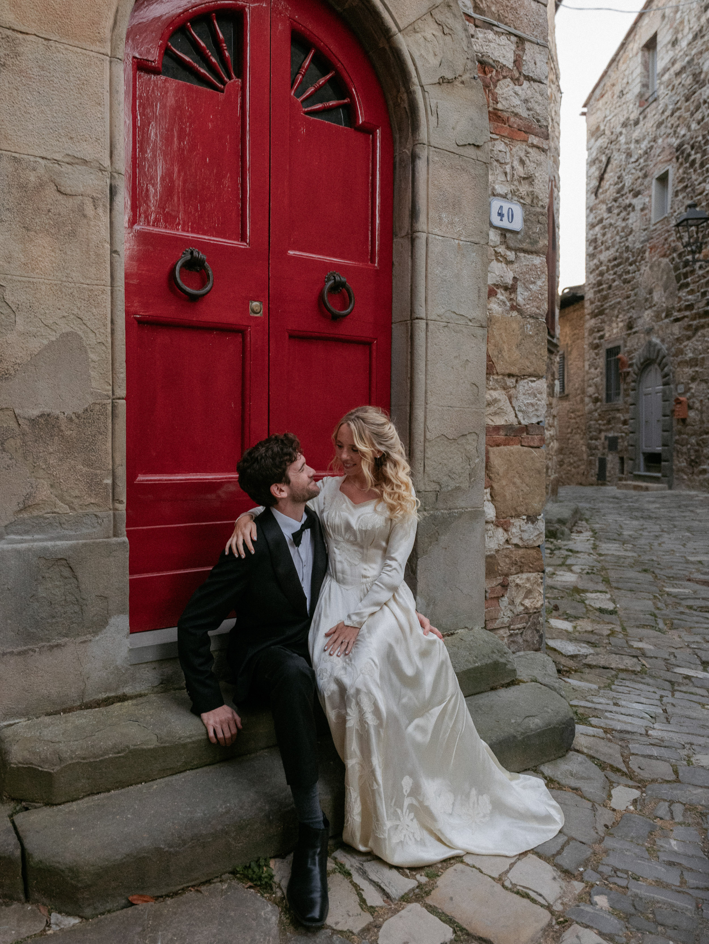 Romantic Tuscan Elopement on Film 327 - An Original 1940s Vintage Dress for Emily & Ned's Intimate Elopement in Tuscany Bride in an original 1940's vintage silk wedding dress sat on a step by a large curved red door on the streets of an Italian town for their Tuscan elopement.