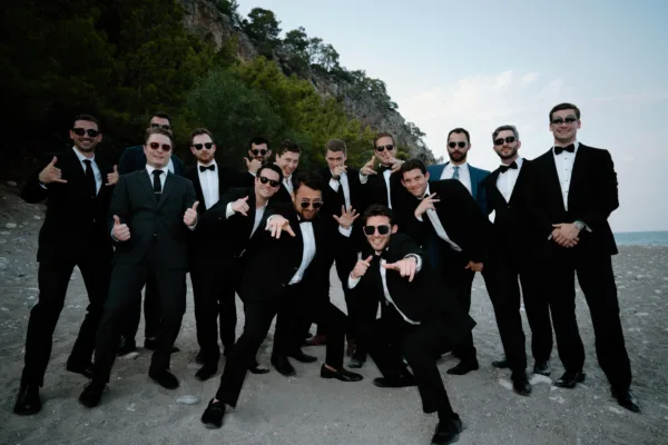 Groomsmen in black tuxedos posing on beach with rocky cliffs at destination wedding in Antalya Turkey by Ramarossi