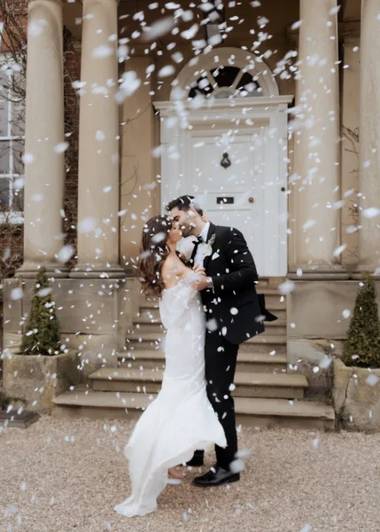 Bride and groom kissing beneath falling confetti outside the grand columned entrance of Iscoyd Park in Shropshire, captured in a soft editorial wedding photography style by Cleo Stella Photography.