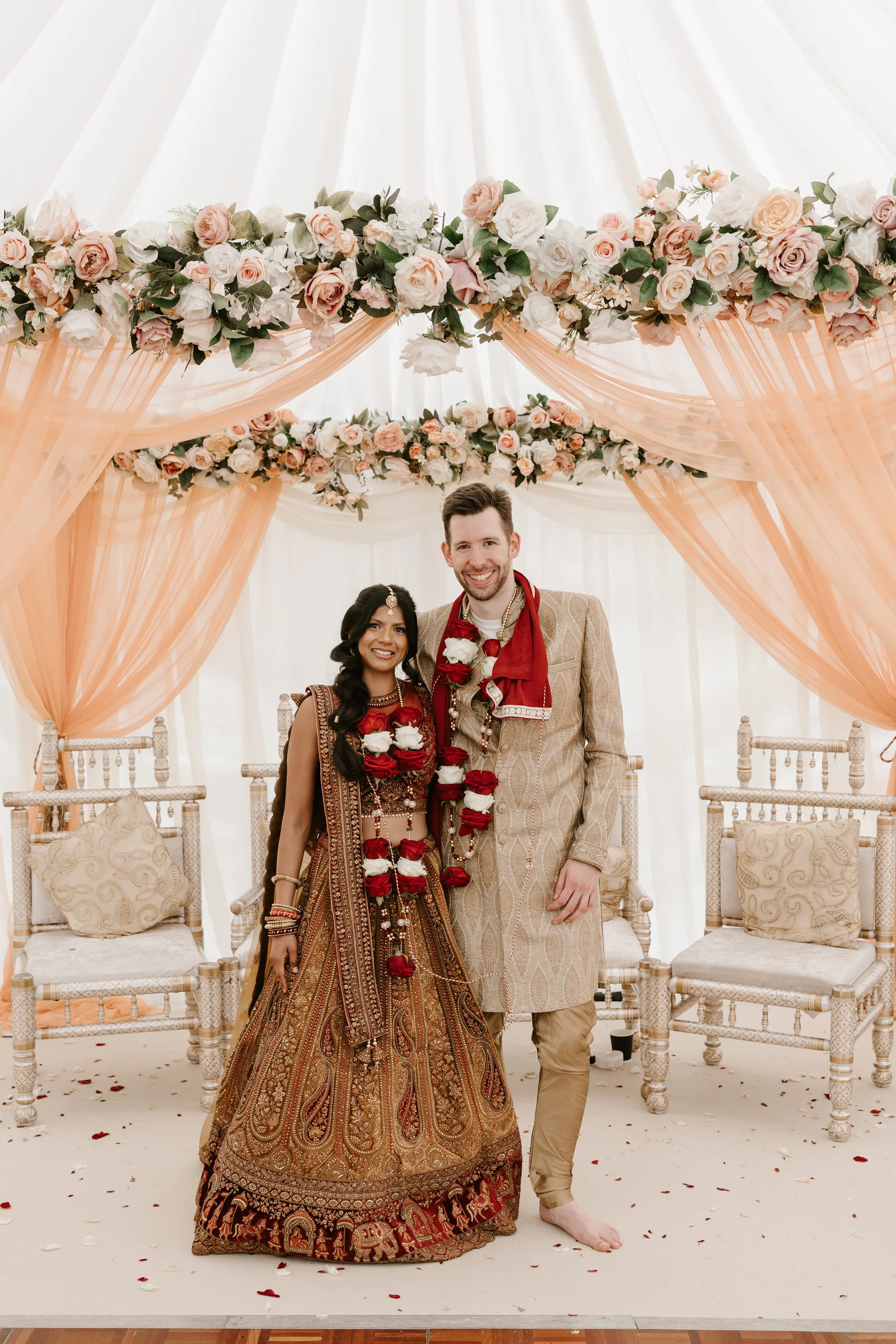 Fusion HIndu Wedding Newburgh Priory 41 - Hema & Will's Fusion Wedding & Hindu Ceremony at Newburgh Priory Hindu wedding ceremony at Newburgh Priory. Bride and groom stand beneath their mandap draped with peach tulle and decorated with peach flowers. Bride wears a read and gold lehenga and groom wears traditional gold Hindu wedding attire. They also wear floral garlands around their necks.