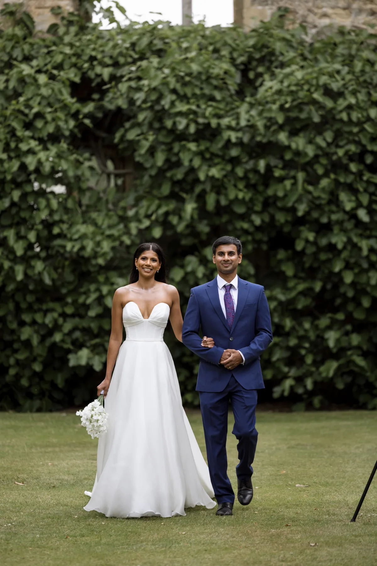 Fusion HIndu Wedding Newburgh Priory 58 - Hema & Will's Fusion Wedding & Hindu Ceremony at Newburgh Priory Brother of the bride in an elegant navy blue suit accompanies his sister towards the outdoors ceremony at Newburgh Priory. Bride clutches a small white bouquet and weras Suzanne Neville.