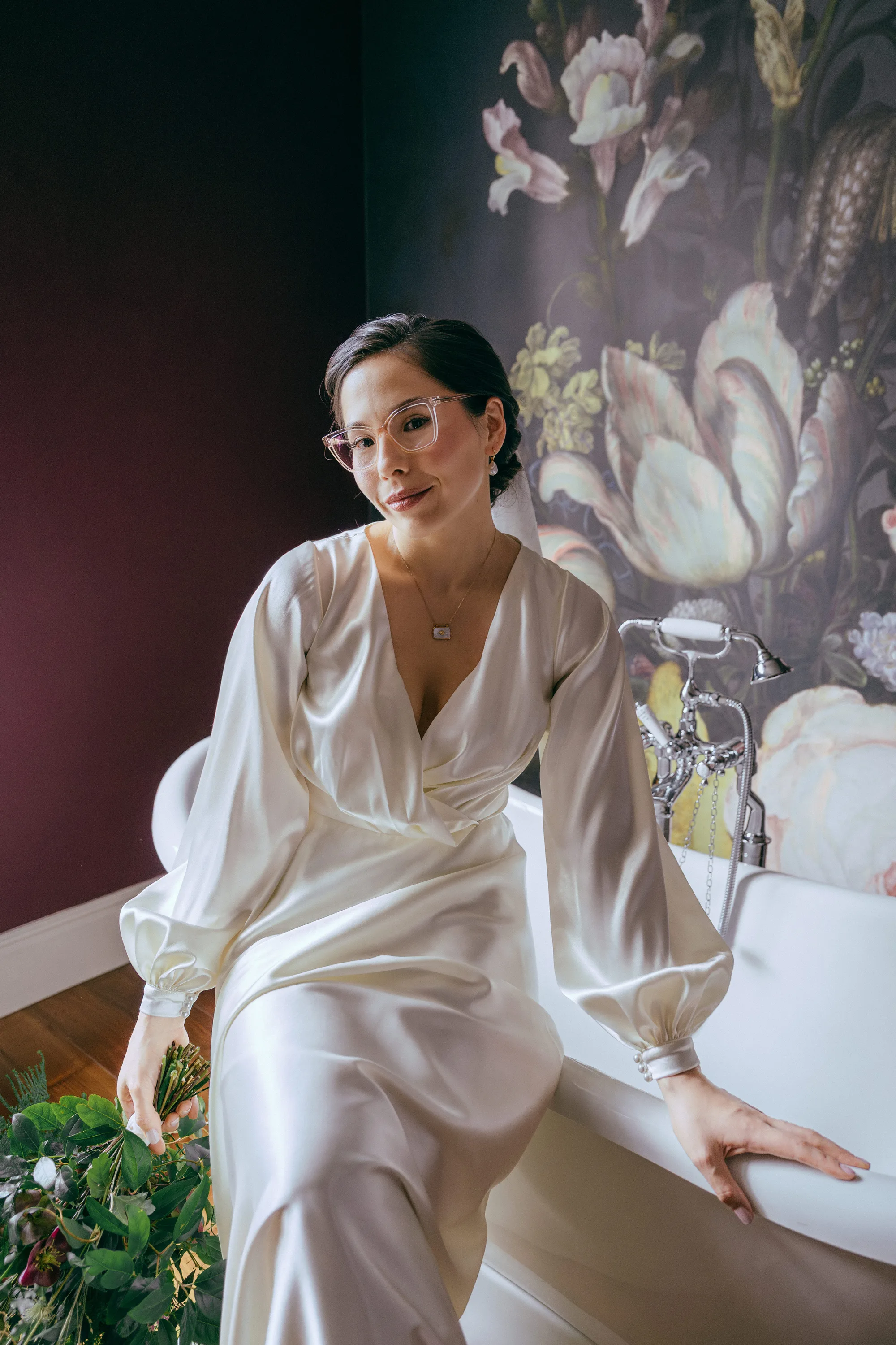 Garthmyl Hall Winter Wedding 63 - Tom & Cami's Heartfelt & Bilingual Winter Wedding at Garthmyl Hall, Wales Bride wearing glasses sat on a roll top bath at Garthmyl Hall in Wales. She wears a silk wedding dress by Lutien Bridal Gowns and carries a green bouquet.