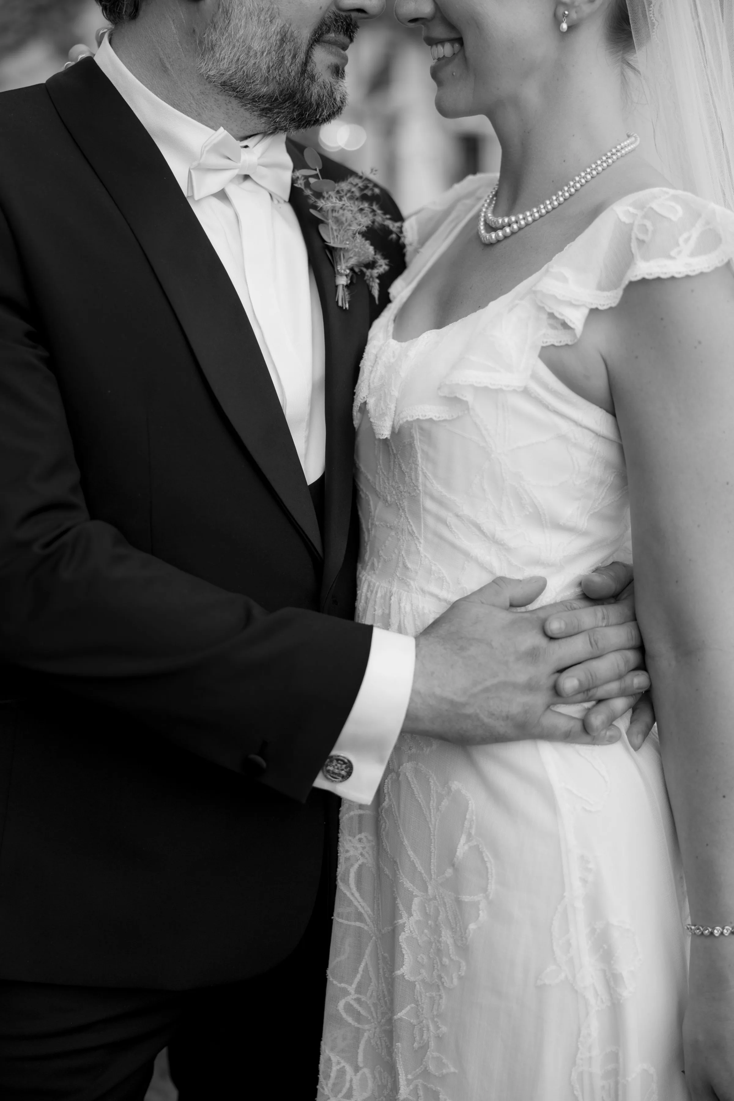 Indie Bride London Chateau Cazenac wedding France 143 - Laura & Ben's Romantic & Intimate Wedding Day Gathering at Chateau Cazenac in The Dordogne Close up shot of bride and groom's torso as groom hugs his bride around the waist. The bride wears a pearl necklace and simple wedding dress with ruffle detail around the neck.