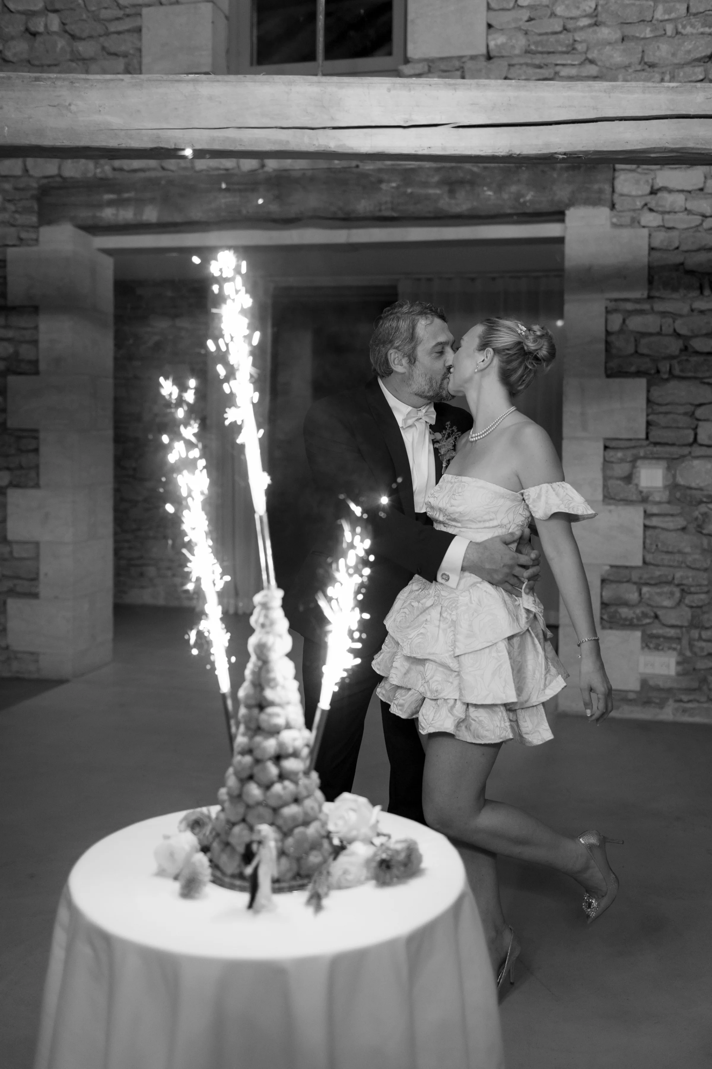 Indie Bride London Chateau Cazenac wedding France 176 - Laura & Ben's Romantic & Intimate Wedding Day Gathering at Chateau Cazenac in The Dordogne Croquembouche tower with sparklers sat on a small round cake table. Behind, the bride and groom embrace one another, the bride wears a cute tiered mini dress to dance and celebrate in.