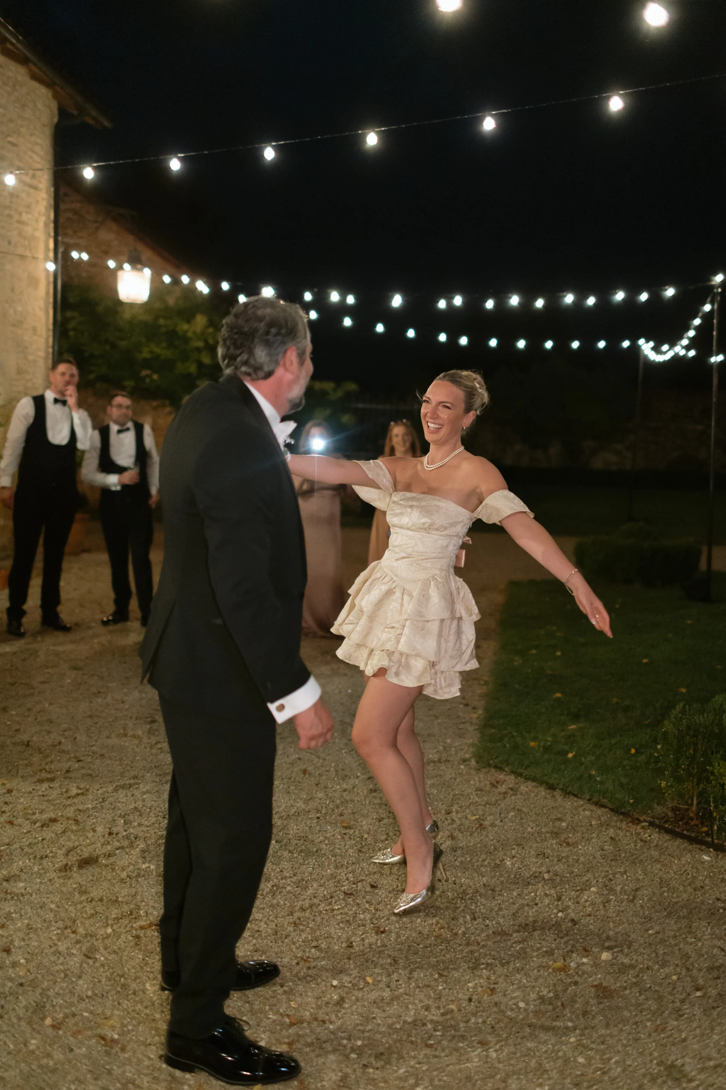 Indie Bride London Chateau Cazenac wedding France 184 1 - Laura & Ben's Romantic & Intimate Wedding Day Gathering at Chateau Cazenac in The Dordogne Bride in a cute, short tiered mini dress dancing with her new husband during the evening of their wedding at Chateau Cazenac. Festoon lights hang above them to light the scene.