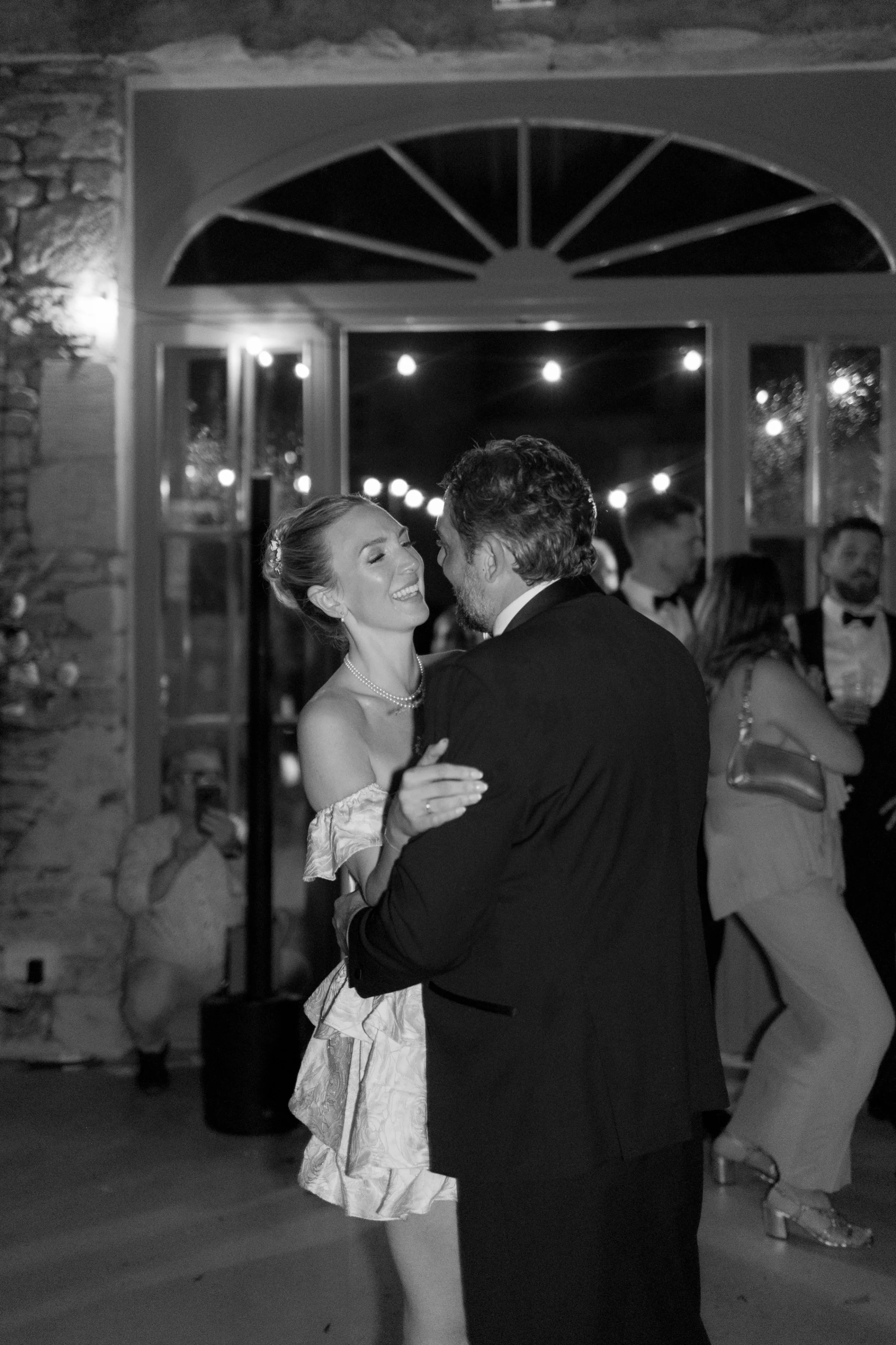 Indie Bride London Chateau Cazenac wedding France 189 - Laura & Ben's Romantic & Intimate Wedding Day Gathering at Chateau Cazenac in The Dordogne Bride in a cute, short tiered mini dress dancing with her new husband during the evening of their wedding at Chateau Cazenac. Festoon lights hang above them to light the scene.