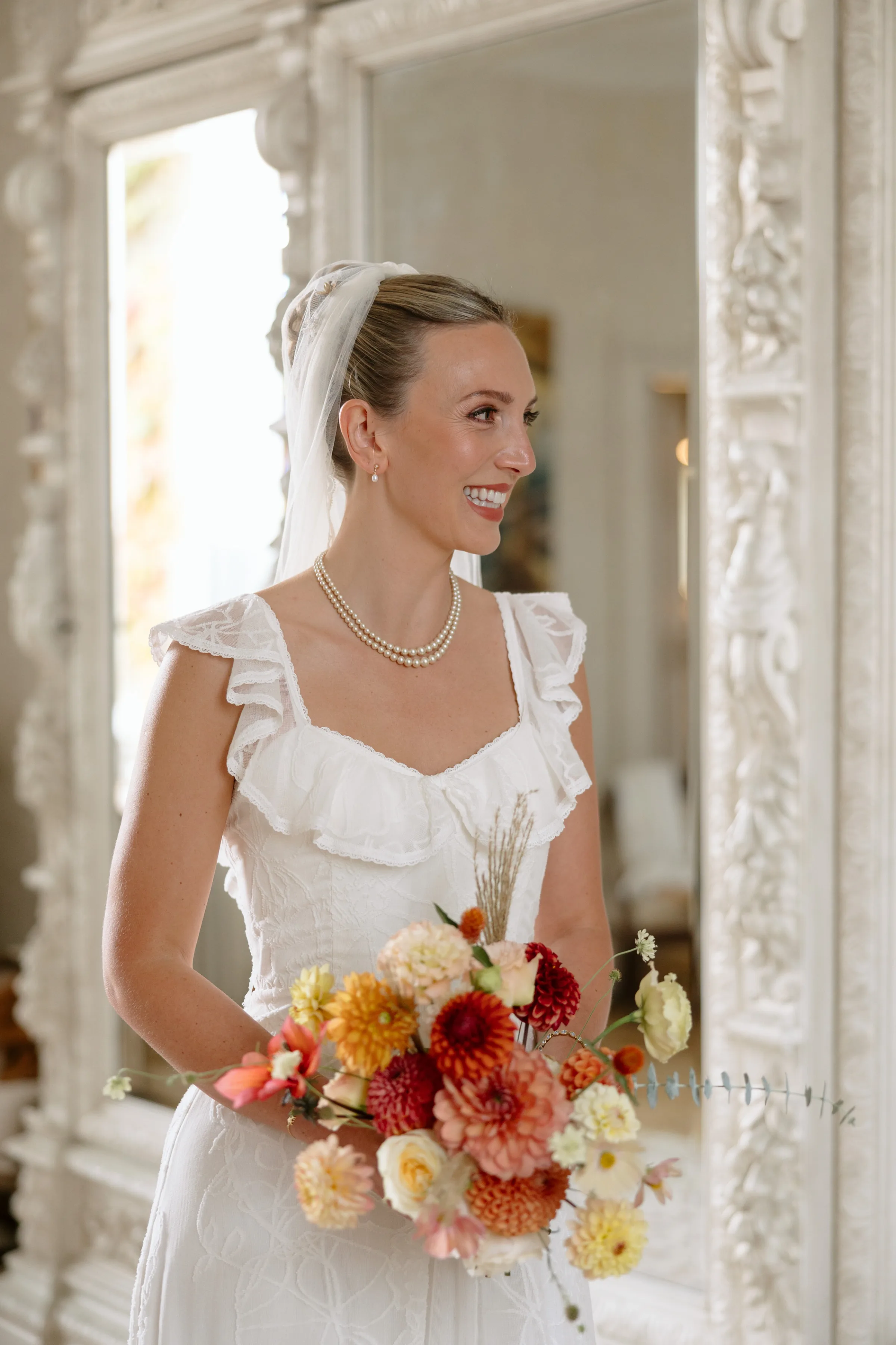 Indie Bride London Chateau Cazenac wedding France 38 - Laura & Ben's Romantic & Intimate Wedding Day Gathering at Chateau Cazenac in The Dordogne Bride wearing her hair up in a high bun, single tier veil and frilly sleeved dress smiles as she stands holding a colourful bouquet of Dahlia in Autumnal shades.