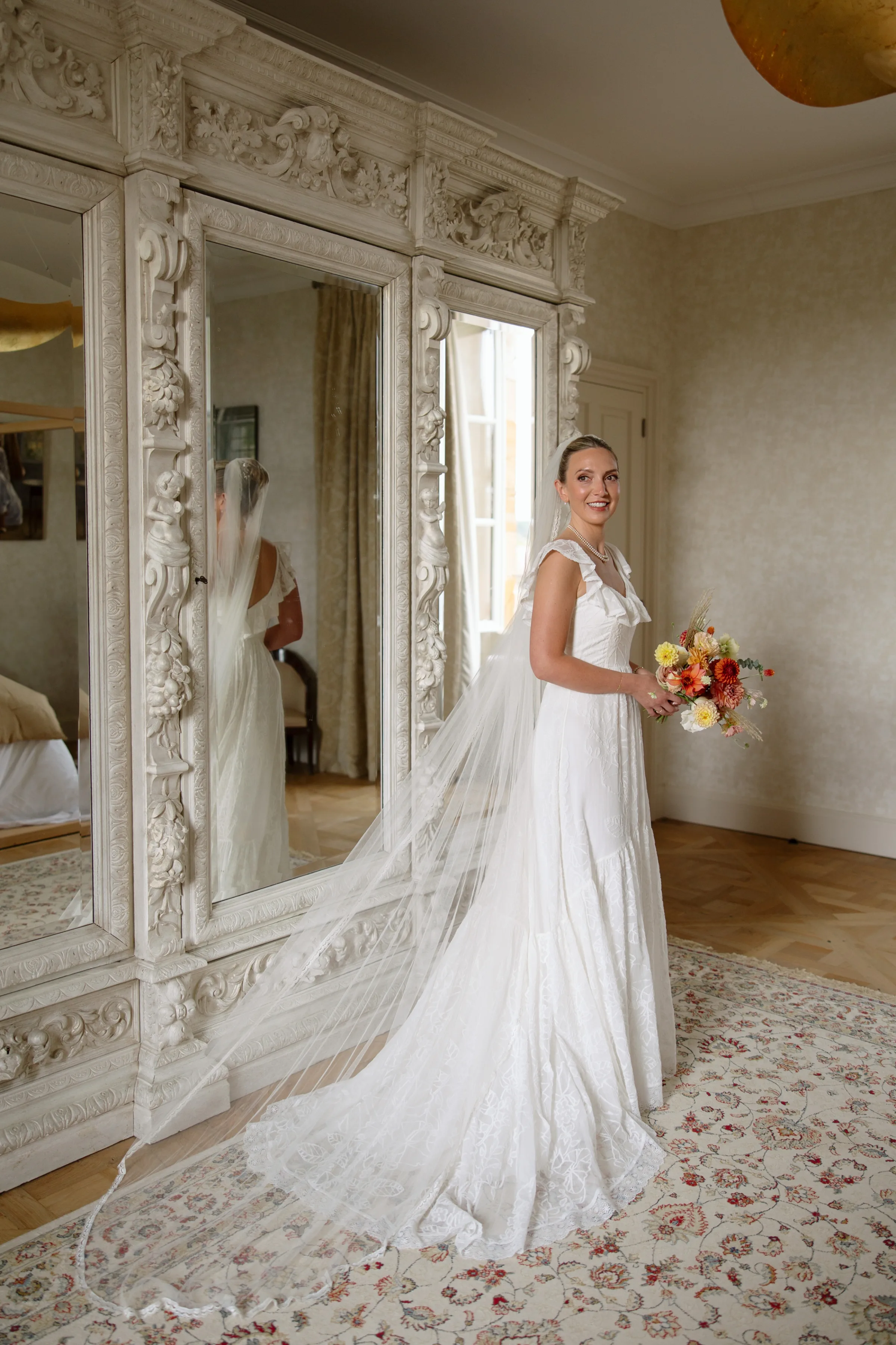 Indie Bride London Chateau Cazenac wedding France 39 - Laura & Ben's Romantic & Intimate Wedding Day Gathering at Chateau Cazenac in The Dordogne Bride wearing her hair up in a high bun, single tier veil and frilly sleeved dress smiles as she stands next to a full length mirror holding a colourful bouquet of Dahlia in Autumnal shades.
