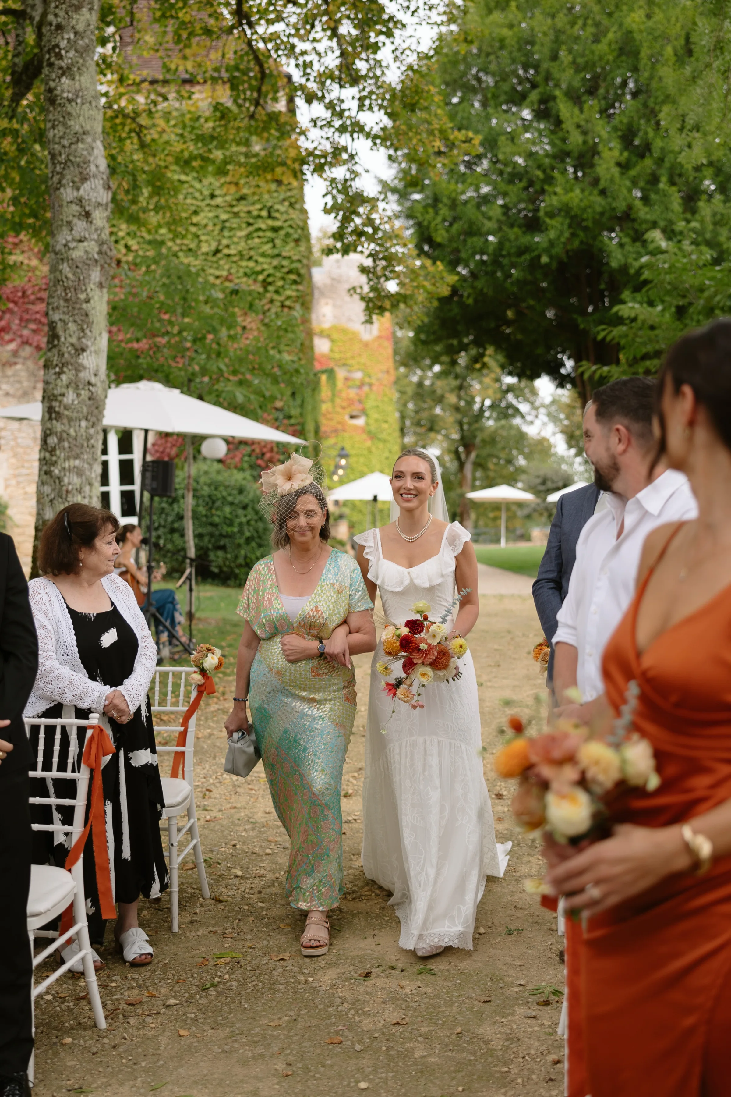 Indie Bride London Chateau Cazenac wedding France 48 - Laura & Ben's Romantic & Intimate Wedding Day Gathering at Chateau Cazenac in The Dordogne Elegant bride wearing a simple wedding dress with ruffle detail around the neck and sleeve is being accompanied by her mother down the aisle for her outdoor wedding at Chateau Cazenac in the Dordogne.