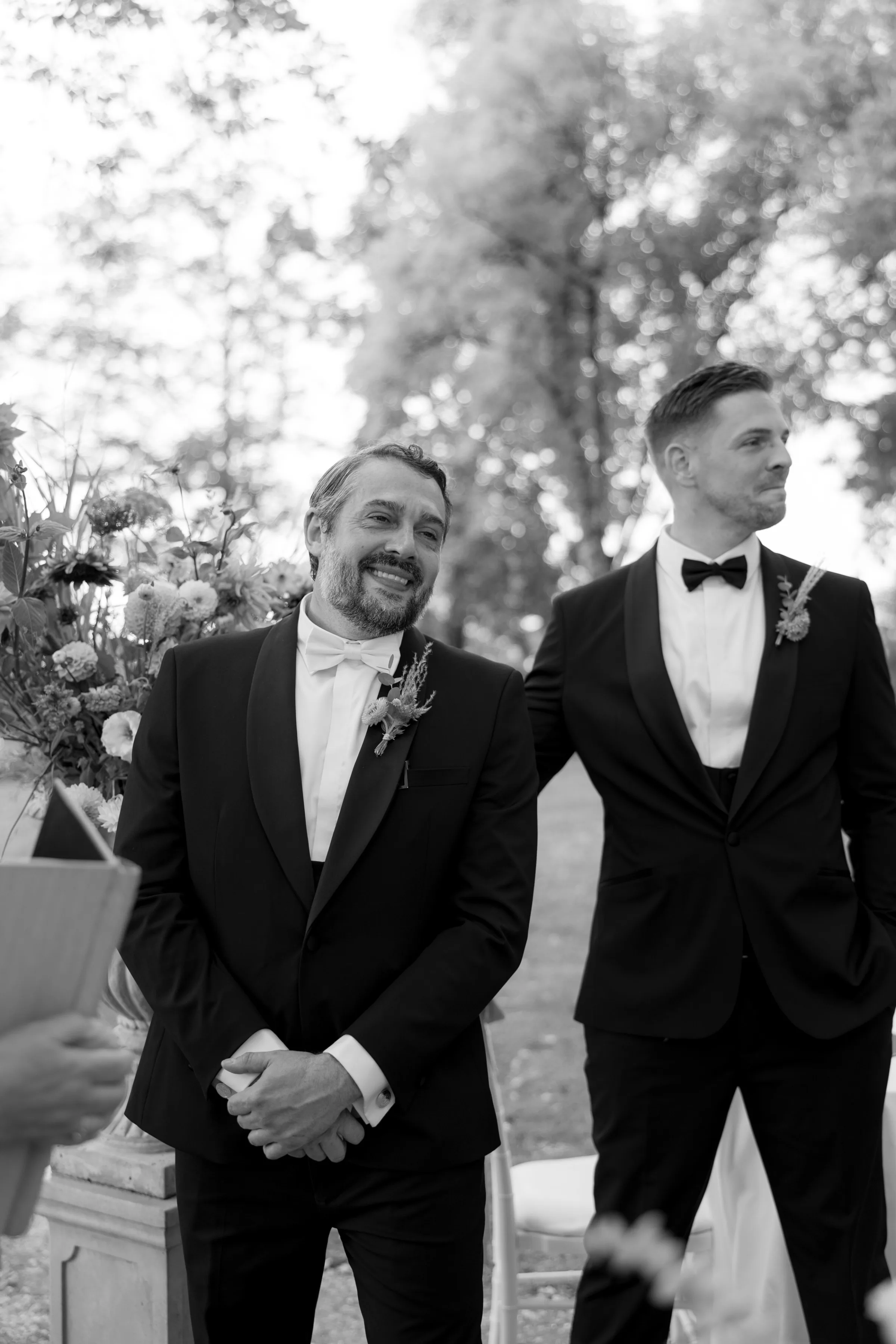 Indie Bride London Chateau Cazenac wedding France 52 - Laura & Ben's Romantic & Intimate Wedding Day Gathering at Chateau Cazenac in The Dordogne Proud groom and emotional best man turn to look as the bride arrives at the ceremony. They wear black tie attire, the groom wears a white bow tie.