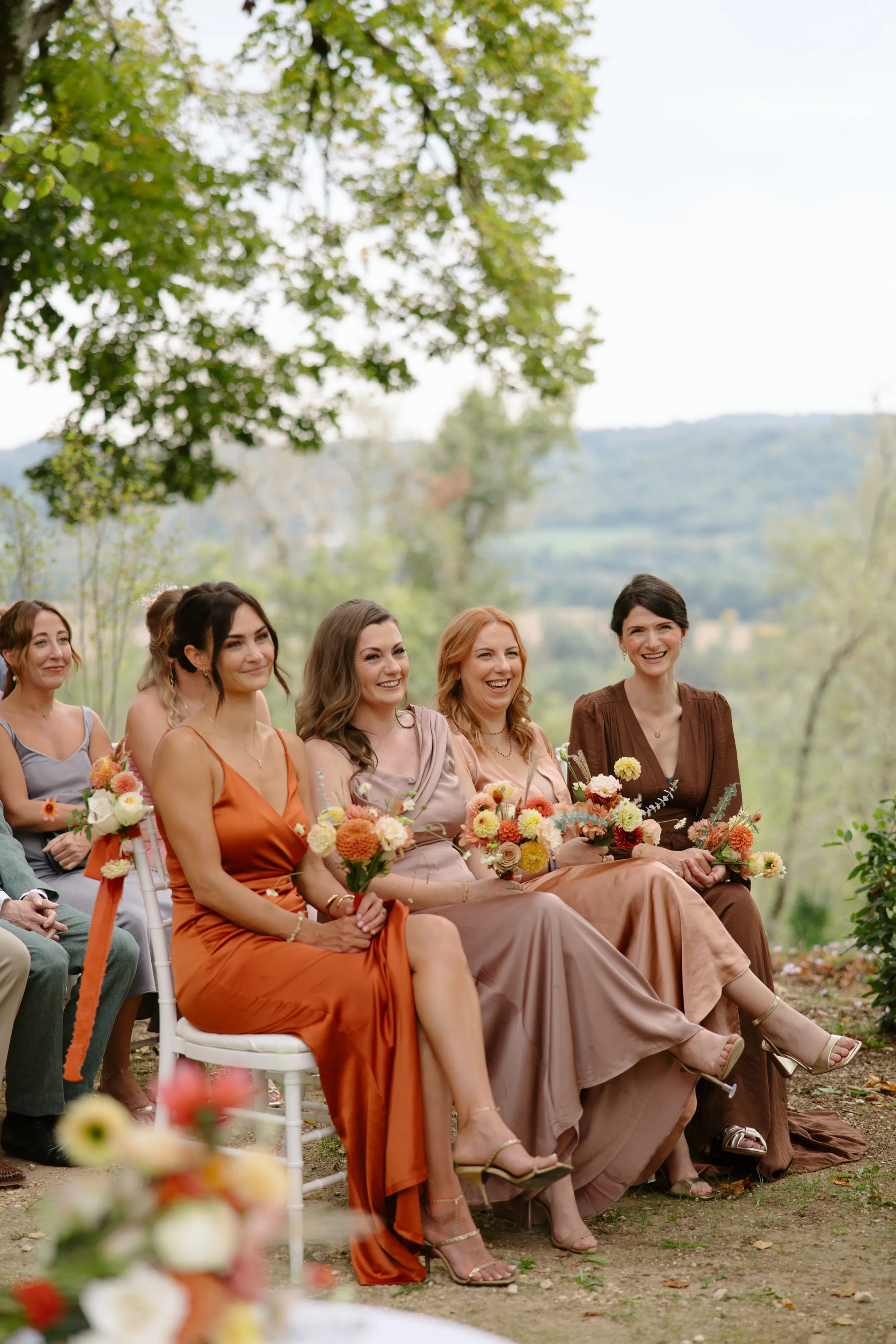 Indie Bride London Chateau Cazenac wedding France 58 1 - Laura & Ben's Romantic & Intimate Wedding Day Gathering at Chateau Cazenac in The Dordogne Bridesmaids in Autumn colour wedding dresses seated and smiling during an outdoor wedding ceremony. They carry small bouquets of Autumn wedding flowers.