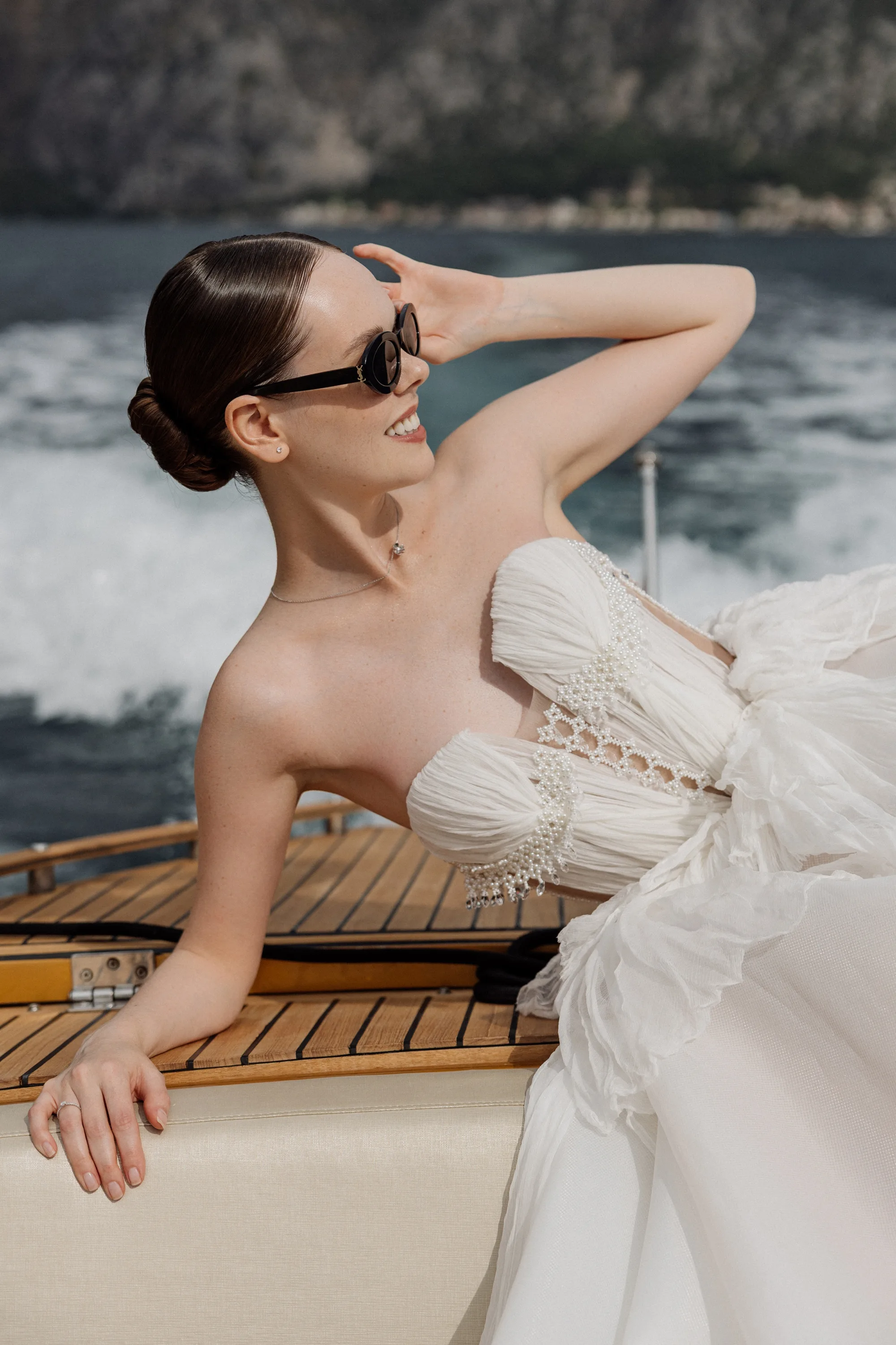 Montenegro Wedding 33 - A Romantic & Intimate Montenegro Wedding at Villa Shah, Inspired by 'La Vita Nobile' Bride leaning back on a speedboat. She wears sunglasses and a Rara Couture gown with compressed silk corset detail. The white waves of a wake created by the boat can be seen behind her.