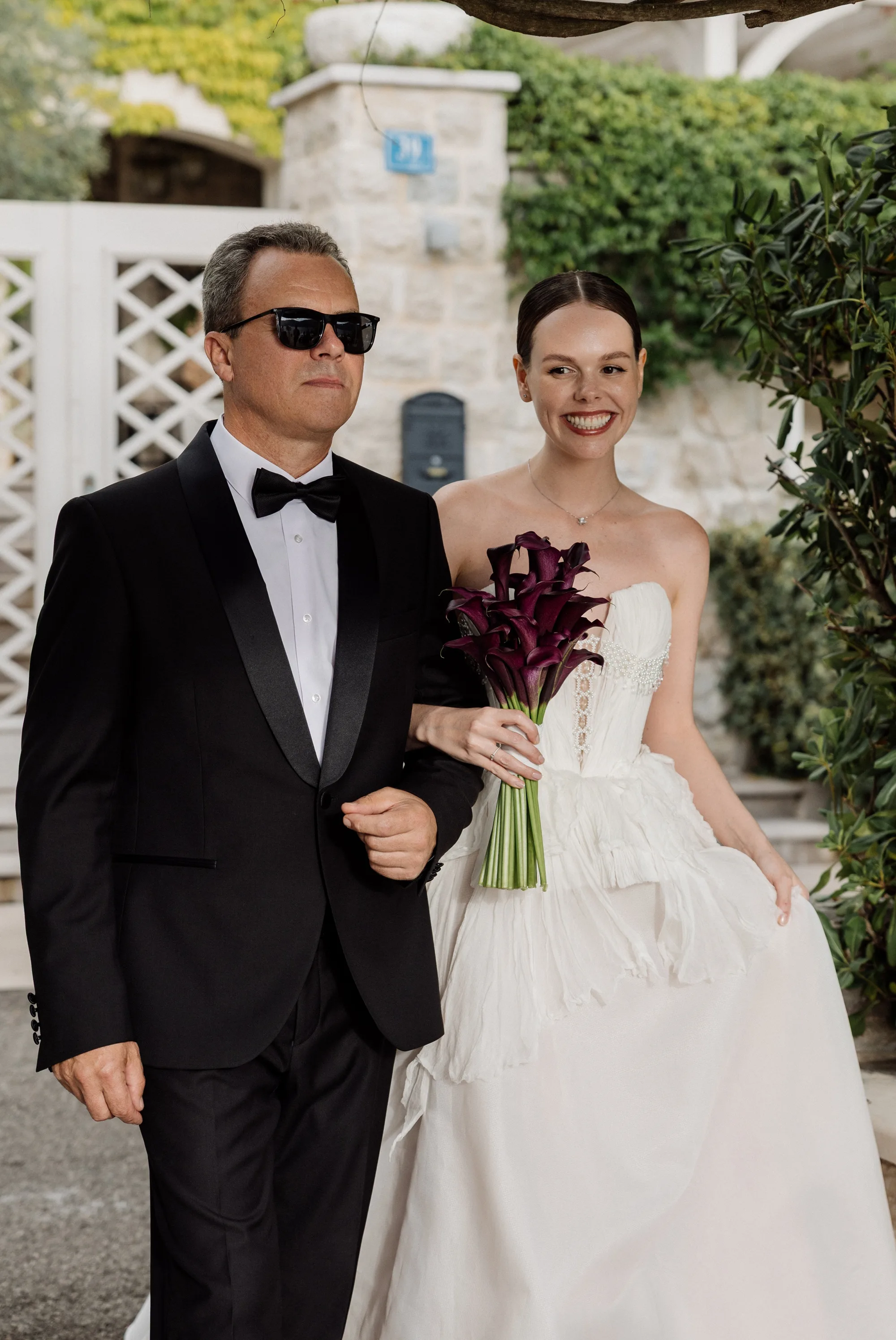 Montenegro Wedding 79 - A Romantic & Intimate Montenegro Wedding at Villa Shah, Inspired by 'La Vita Nobile' Bride being accompanied by her father down the aisle. She carries a bouquet of dark lillies and wears a dress by Rara Avis. Her father is in black tie and sunglasses.
