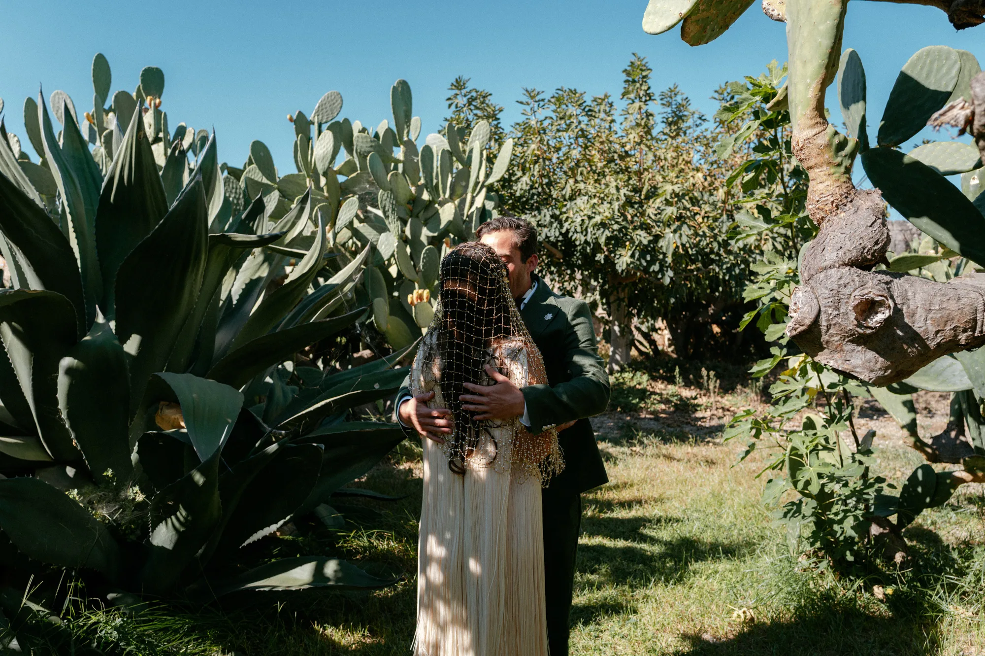 Multiple Day Destination Wedding in Masseria Potenti Puglia Italy 33 - Yael Wore Chana Marelus & A Handmade Beaded Veil for her Relaxed Italian Masseria Wedding Bride wearing a Chana Marelus with tassels and a beaded veil being embraced by her groom in. a green linen suit in the grounds of their Italian wedding venue.