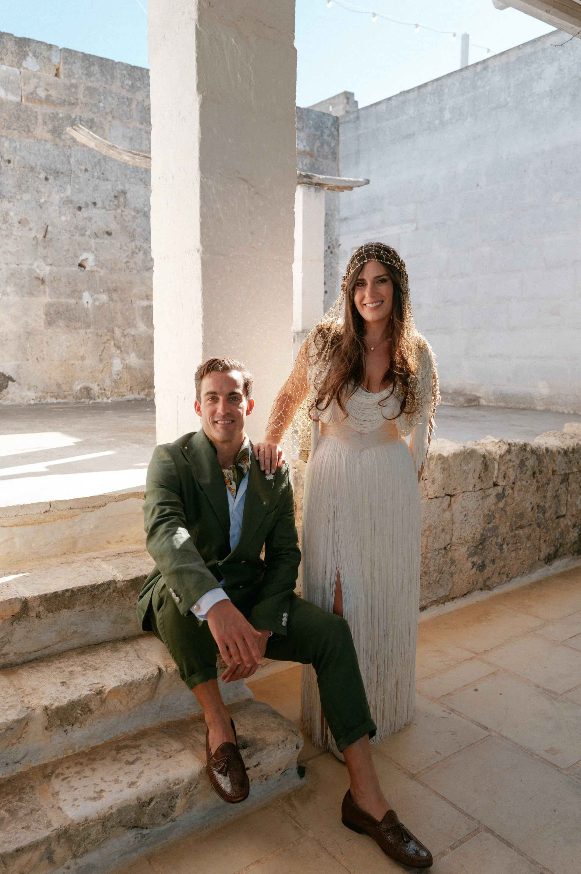 Multiple Day Destination Wedding in Masseria Potenti Puglia Italy 42 - Yael Wore Chana Marelus & A Handmade Beaded Veil for her Relaxed Italian Masseria Wedding Bride wearing a Chana Marelus tassel dress and beaded veil standing by her groom who is seated on a stone step, he wears a green linen suit and cravat.