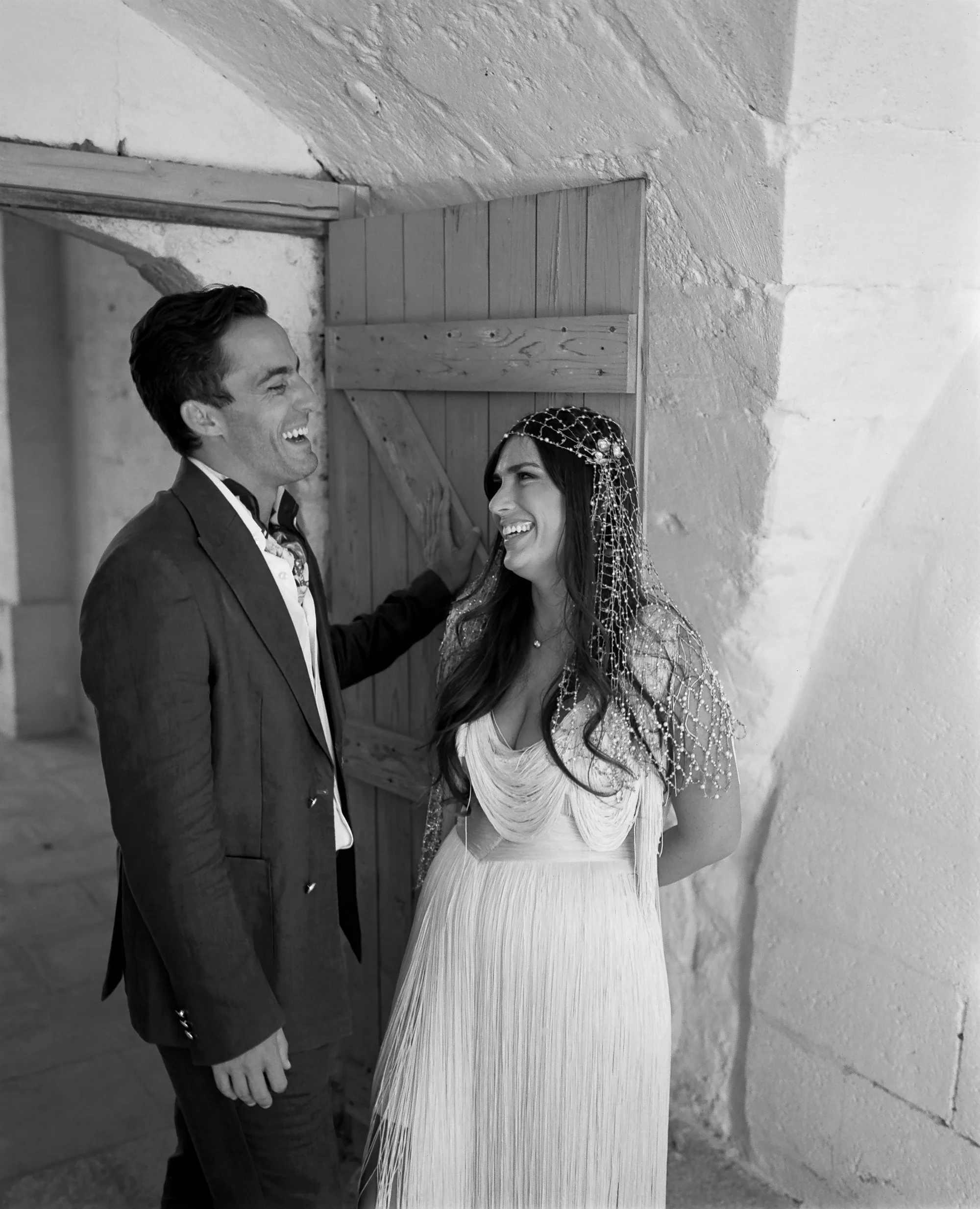 Multiple Day Destination Wedding in Masseria Potenti Puglia Italy 46 - Yael Wore Chana Marelus & A Handmade Beaded Veil for her Relaxed Italian Masseria Wedding Black and white photograph of a bride wearing a Chana Marelus with tassels and a beaded veil, she stands next to her groom in a linen suit and cravat. They are standing in a doorway and laughing.