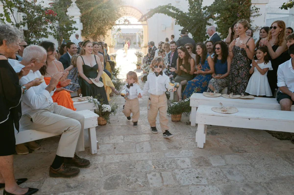 Multiple Day Destination Wedding in Masseria Potenti Puglia Italy 67 - Yael Wore Chana Marelus & A Handmade Beaded Veil for her Relaxed Italian Masseria Wedding Two little ring bearer page boys wearing pale trousers and white shirts and carrying bubble guns as they walk down the aisle.