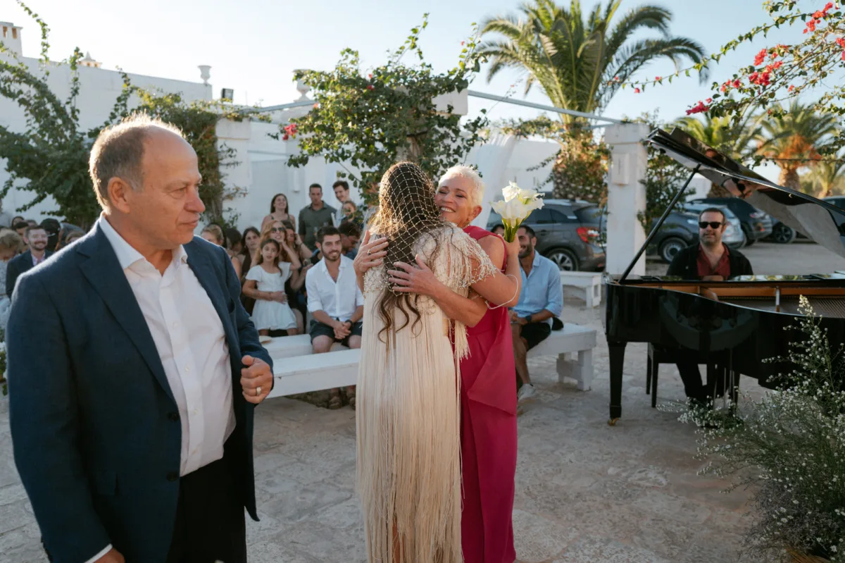 Multiple Day Destination Wedding in Masseria Potenti Puglia Italy 70 - Yael Wore Chana Marelus & A Handmade Beaded Veil for her Relaxed Italian Masseria Wedding Bride in a beaded wedding veil turning to embrace her mother in a red dress as she arrives for her wedding ceremony. Beside them is a pianist playing on a grand piano. Puglia Italy wedding.